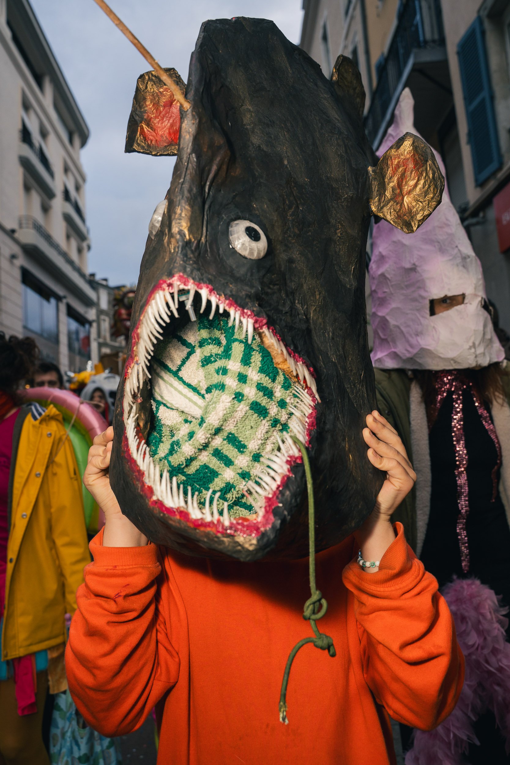 Person wearing a large homemade monster mask with sharp teeth and eyes, holding the mask on their head during a street parade.