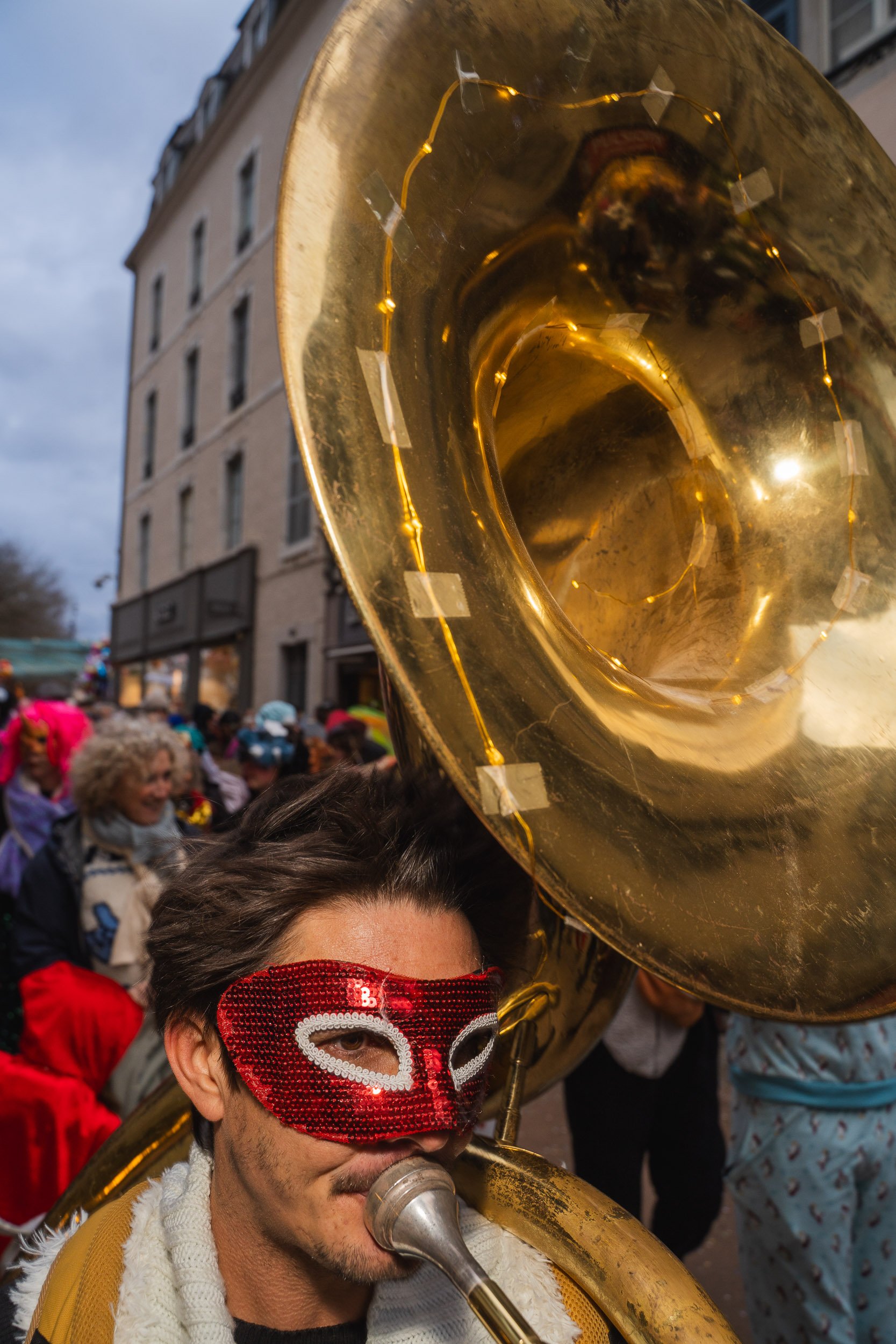 A man wearing a red mask playing a sousaphone during a parade or street event, with other people in costumes and colorful outfits in the background.