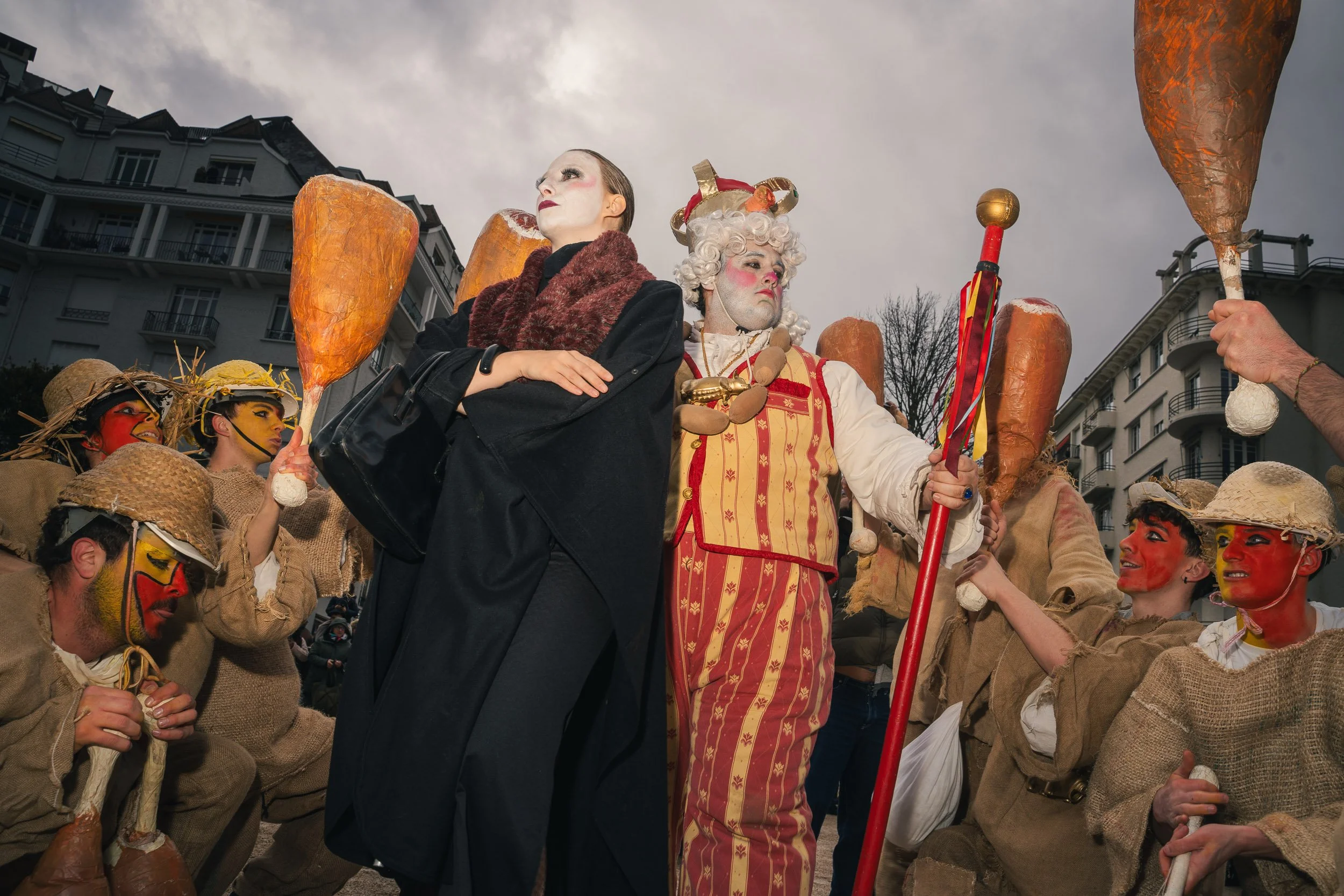 People dressed in theatrical costumes, with some painted faces, gathered outdoors in an urban setting during a parade or festival.