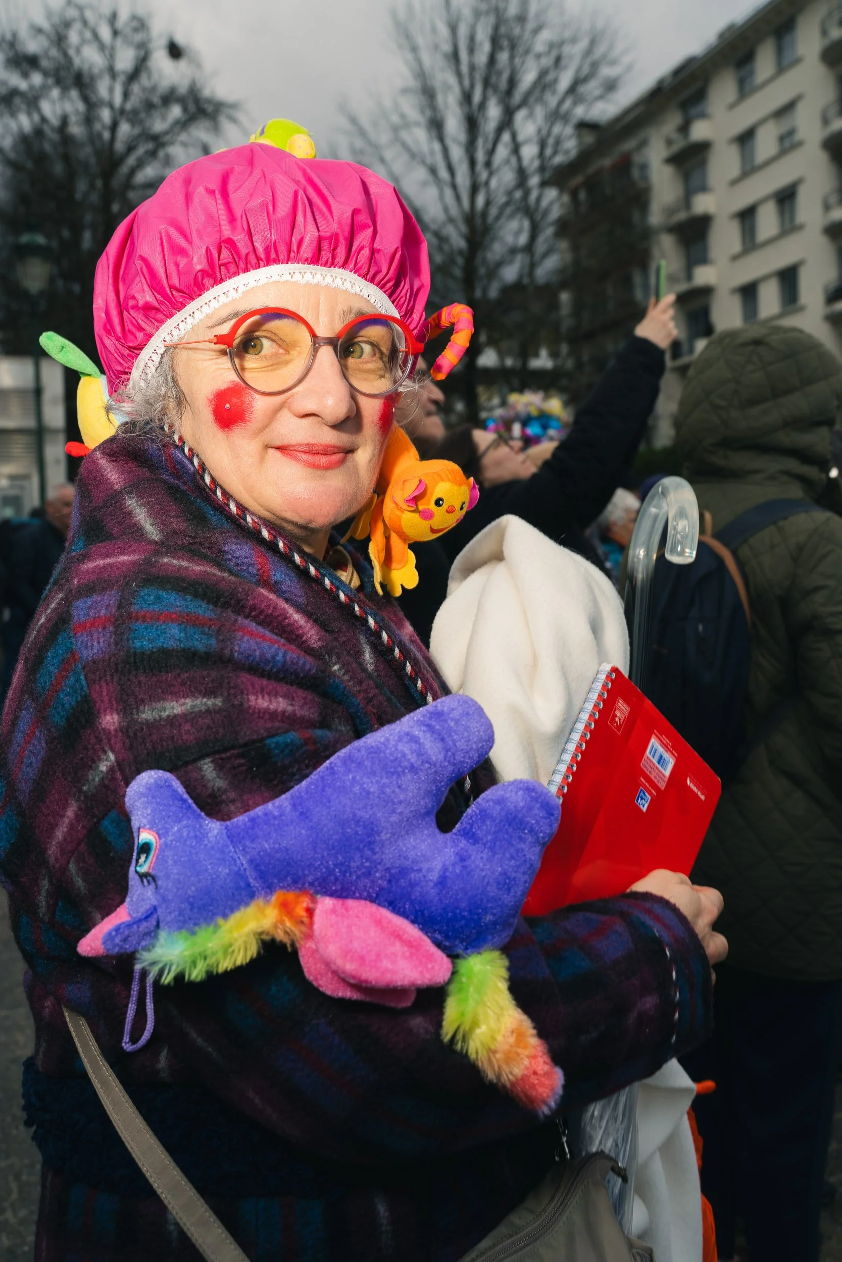 An older woman dressed as a clown, wearing a pink hat, colorful plush accessories, and purple gloves, standing outdoors among a crowd, holding a white hat and a red notebook.