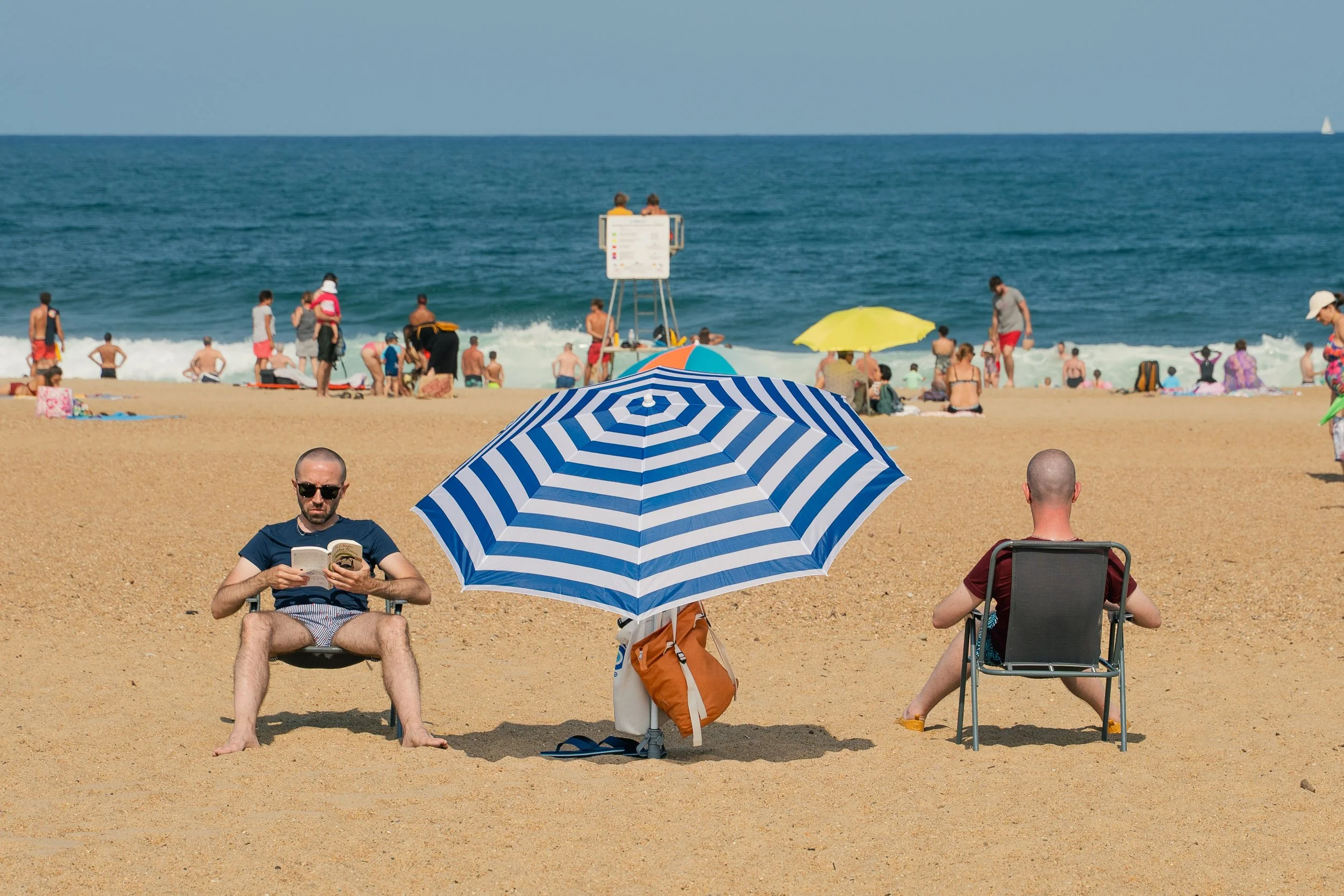People relaxing on a sandy beach under umbrellas, with the ocean in the background, including a man reading a book, another sitting in a lounge chair, and there are many beachgoers near the water.