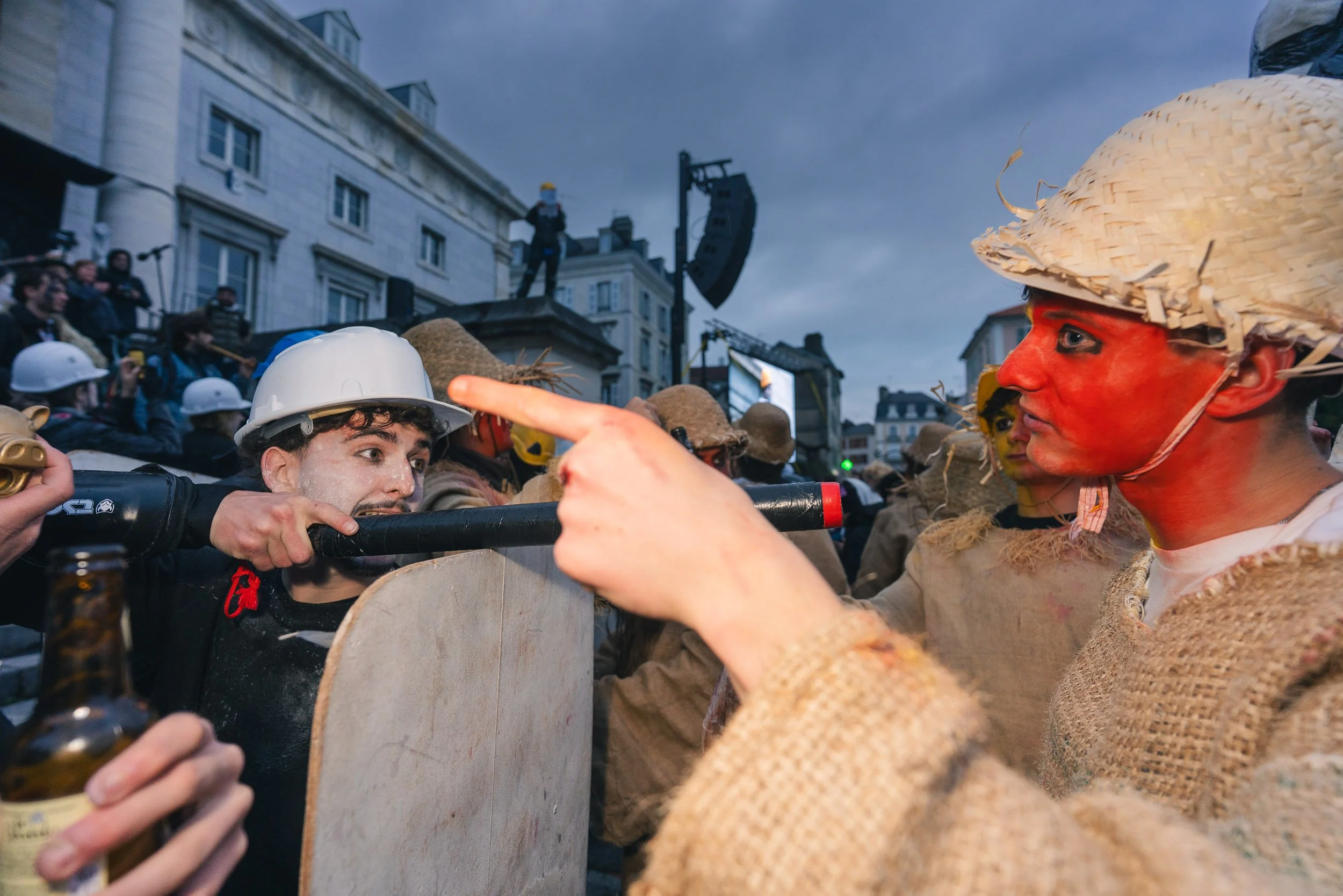 People dressed in costumes and masks, engaging in a protest or street event, with buildings and a stage in the background.