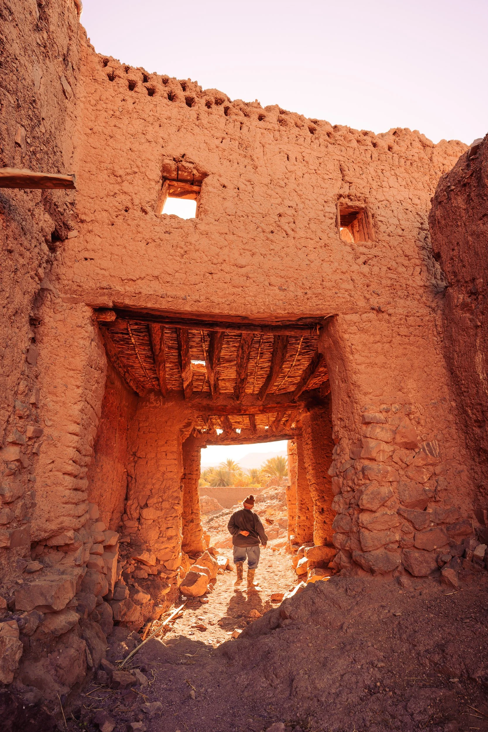 A person standing in front of an ancient, weathered, mud-brick structure with a large arched doorway, two small windows above, and a thatched roof component, set against a desert landscape with palm trees in the background.