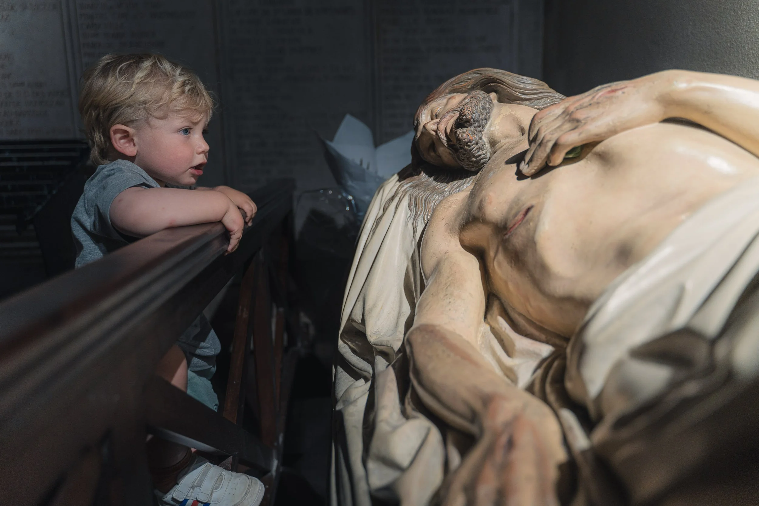 A young child with blonde hair leaning on a wooden railing, looking at a reclining sculpture of Jesus Christ.