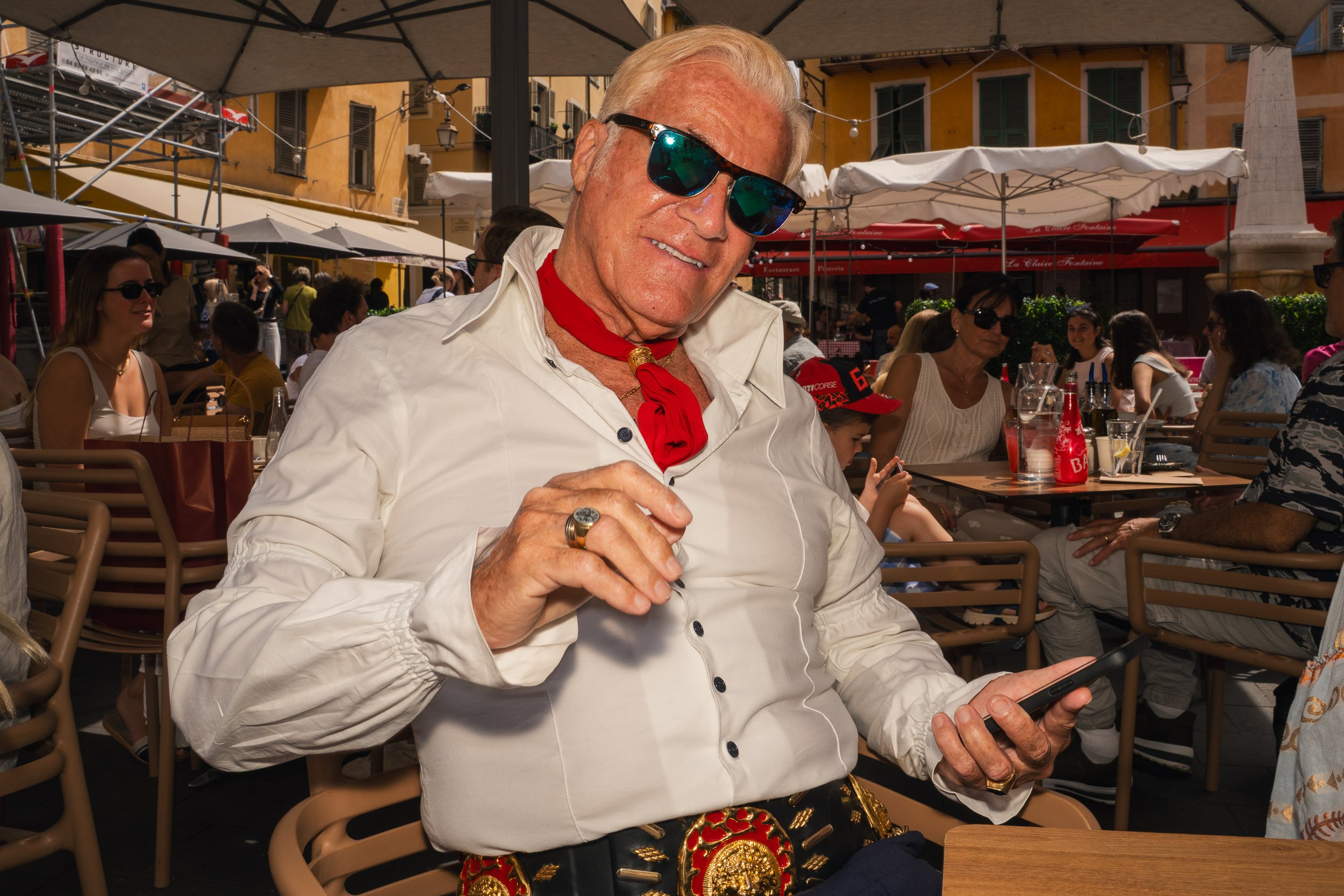An older man with blonde hair wearing sunglasses, a white shirt with rolled-up sleeves, a red neckerchief, and a decorative belt, is sitting at an outdoor restaurant on a sunny day, holding a smartphone in one hand and looking at the camera.