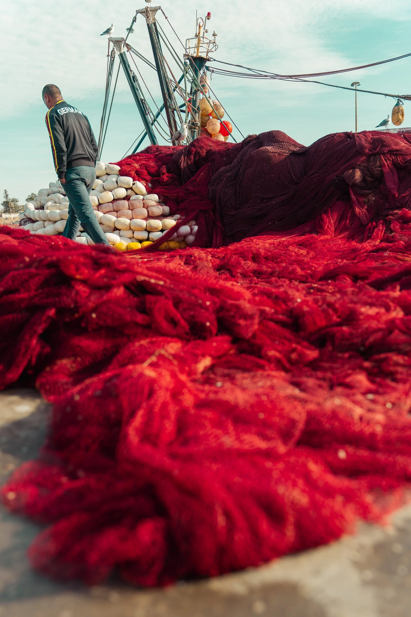 A man walk past large piles of red and black fishing nets on a dock, with fishing buoys and a boat in the background, under a cloudy sky.