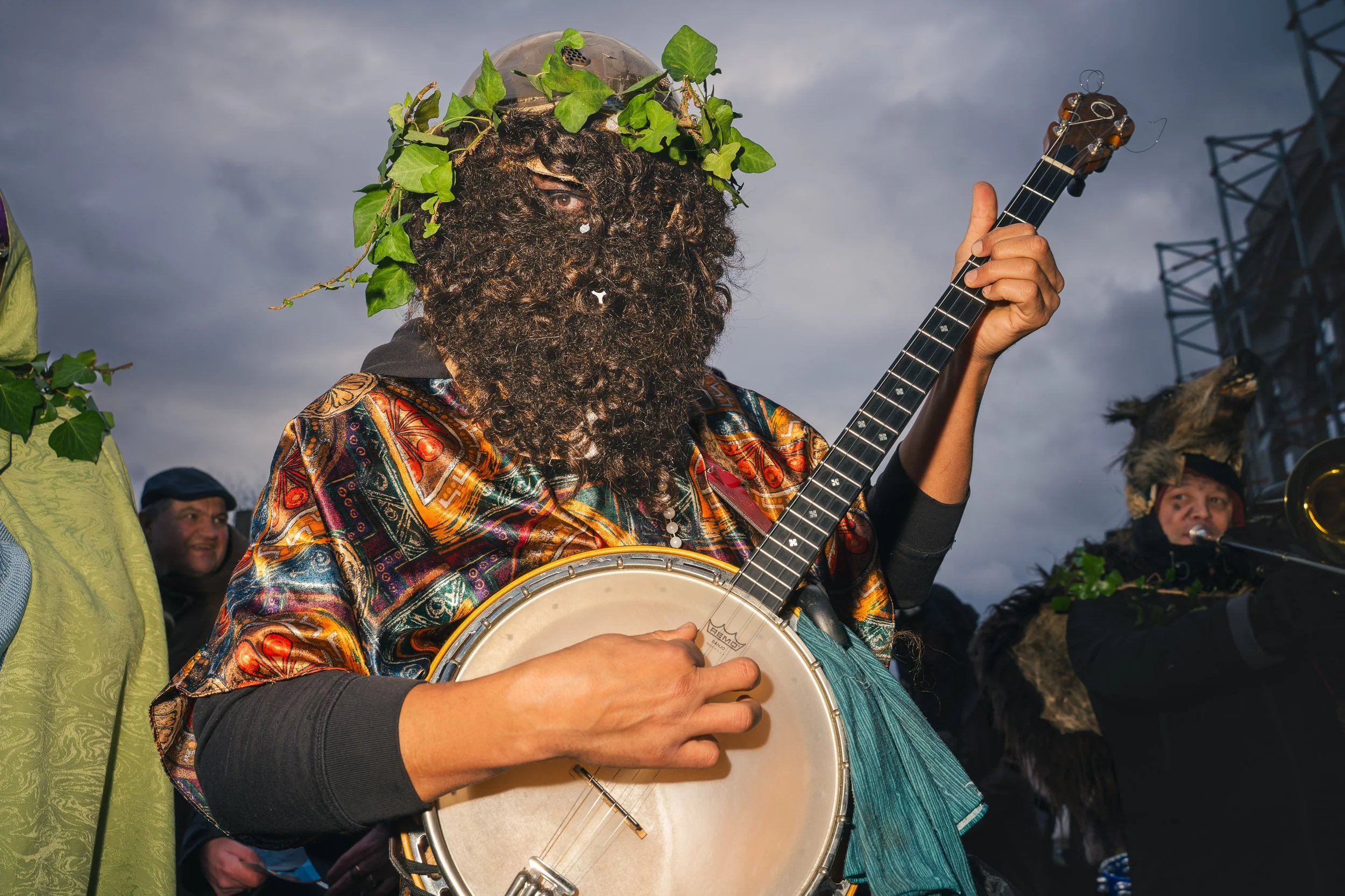 A person with a leafy headpiece and a curly beard playing a banjo, wearing a colorful patterned garment, outdoors during evening with cloudy sky, accompanied by others dressed in costumes.