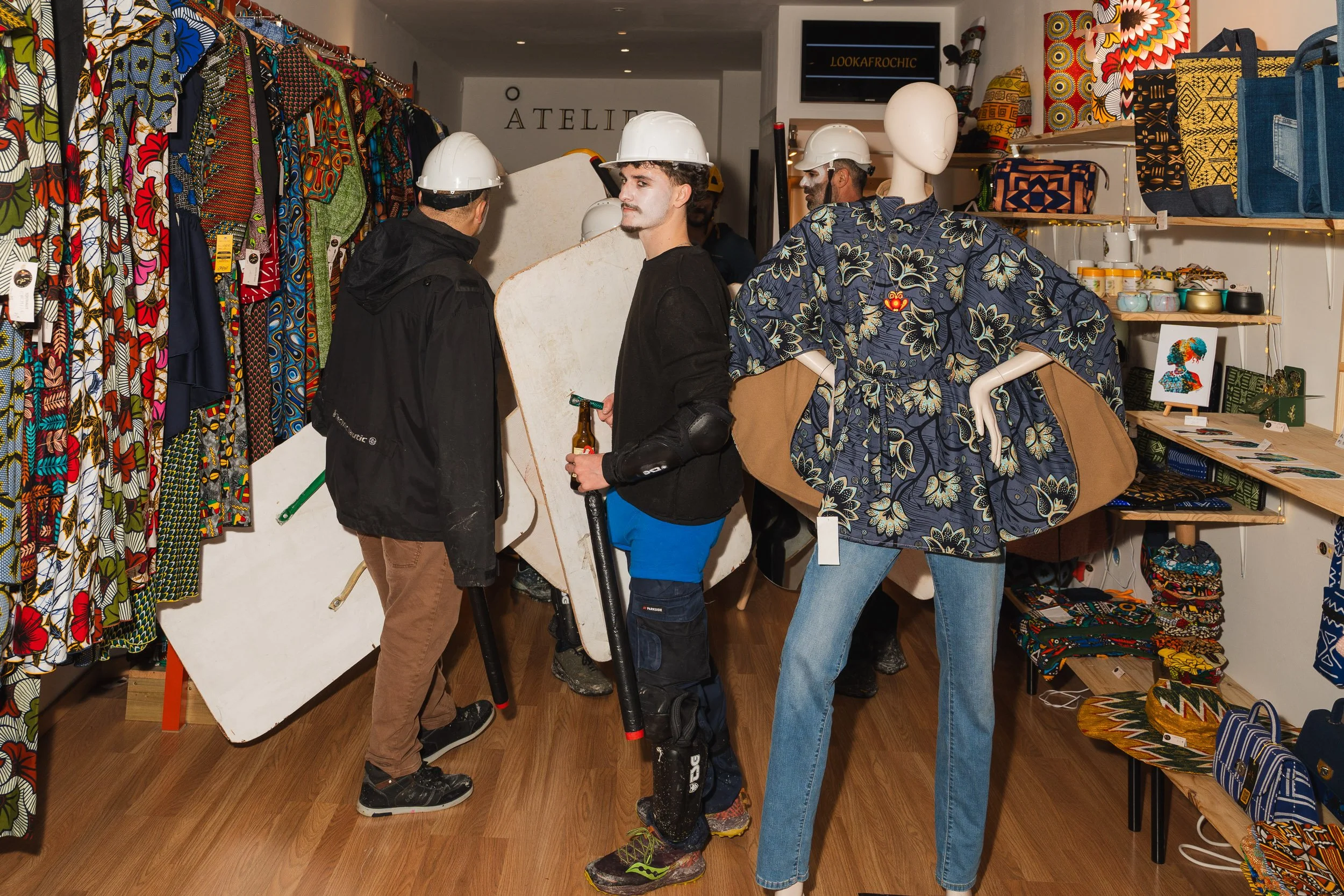 People in a colorful clothing store, some wearing white safety helmets, shopping and browsing among clothing racks, mannequins dressed in patterned garments, and shelves with various accessories.