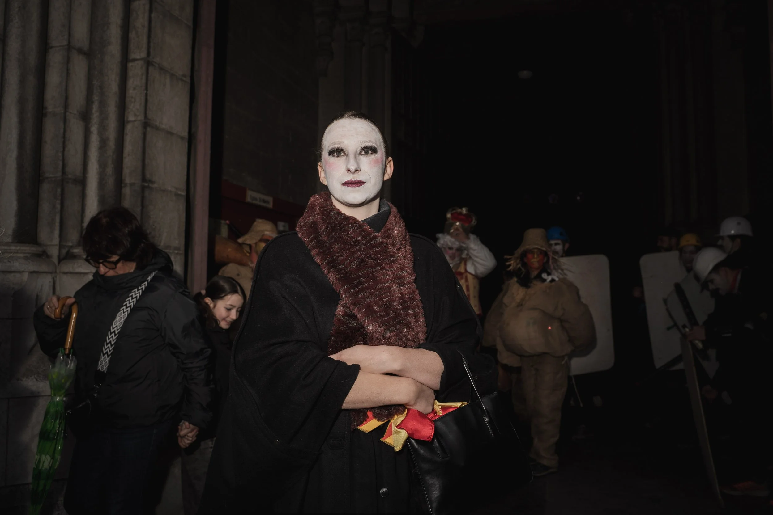 A person with theatrical makeup and dark clothing stands in front of a group of people dressed as clowns and protesters, some wearing helmets and masks, in a dark environment.