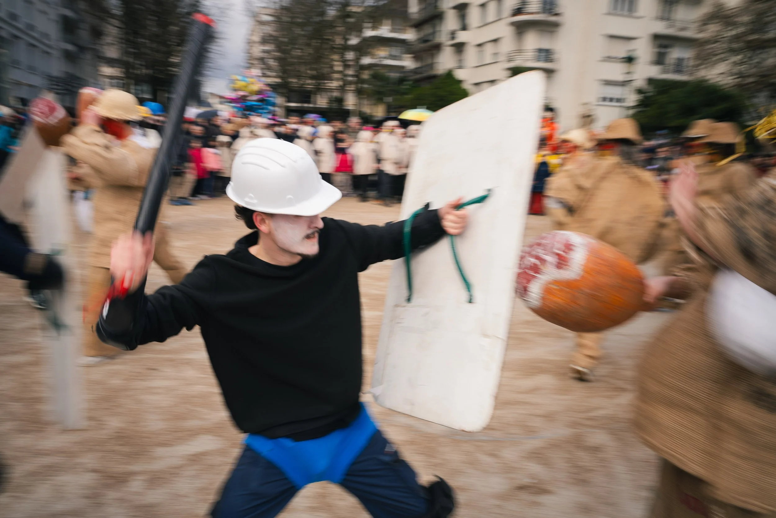 Person dressed as a protestor or riot police, wearing a hard hat, holding a shield and baton, in a chaotic scene with a crowd and buildings in the background.
