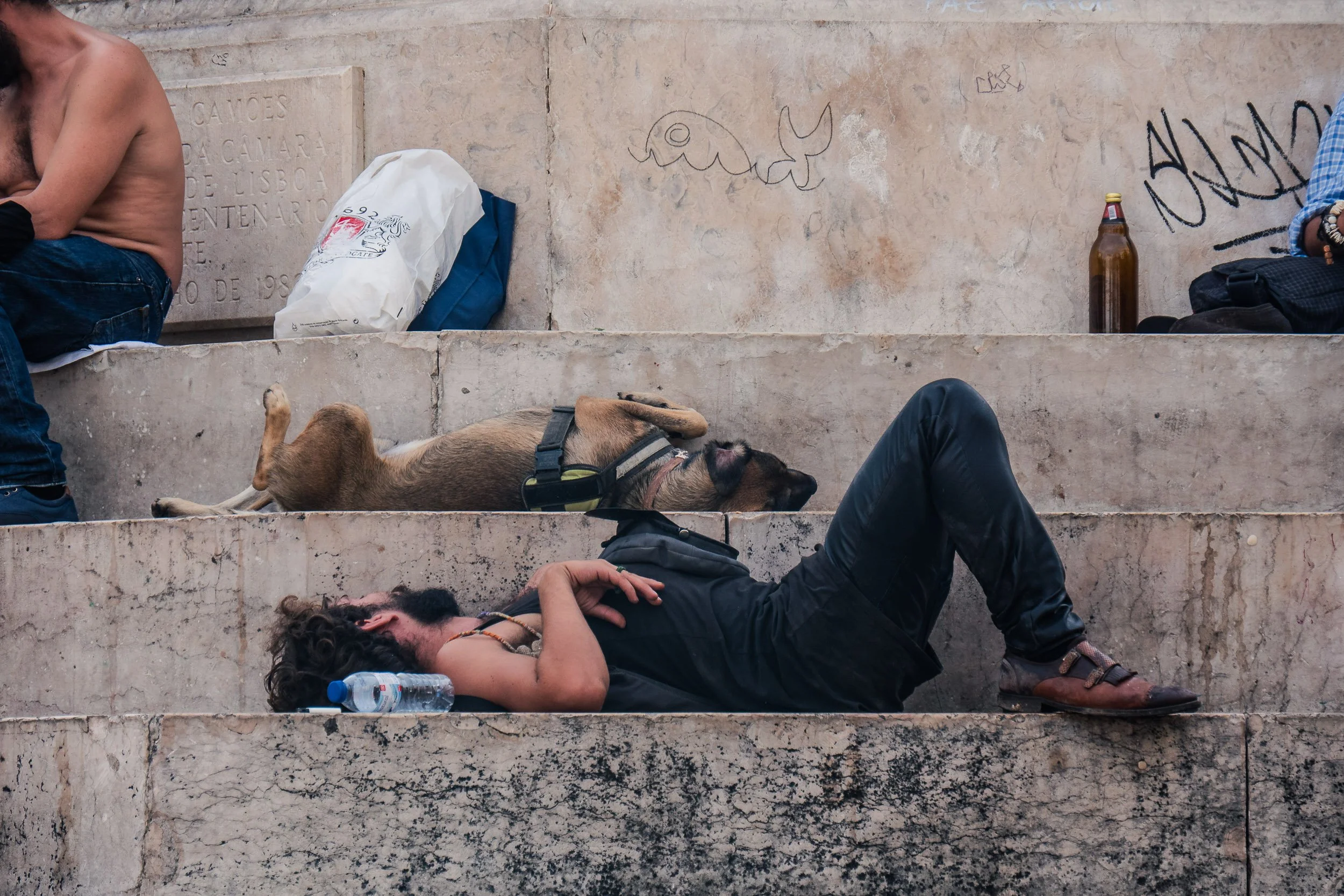A man with long curly hair and a beard, dressed in black, sleeping on stone steps with a water bottle beside him. A dog lies next to him, also sleeping, on the same step. In the background, a person sits shirtless with their arms crossed, and another