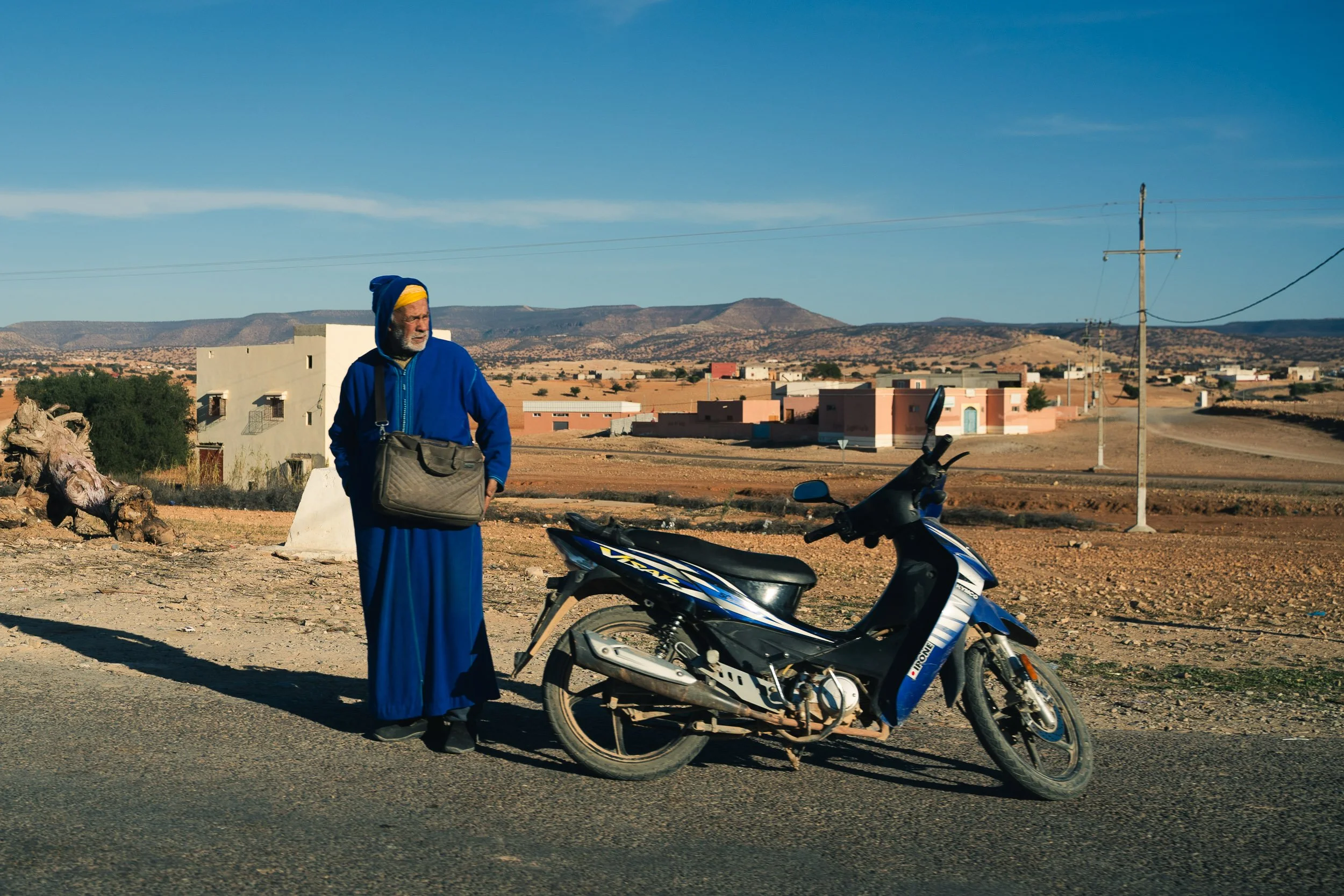 An elderly man dressed in traditional blue attire stands beside a black and blue motorbike on a dry, rural road with a desert landscape, houses in the background, and mountains under a clear blue sky.