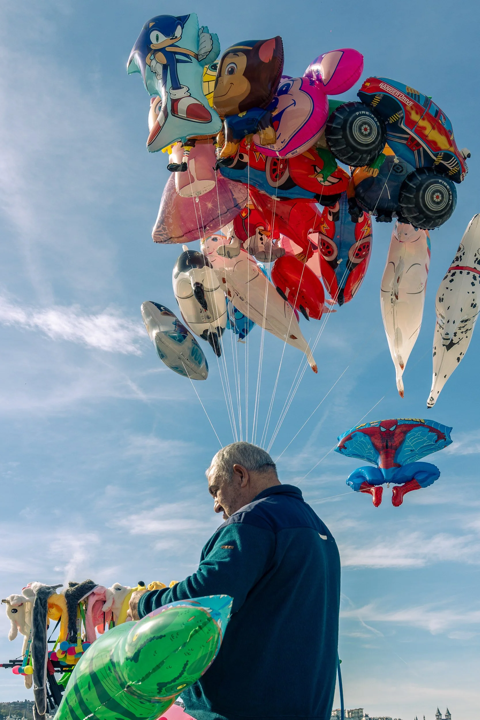 A man at an outdoor market stall with numerous colorful character-shaped and vehicle-shaped helium balloons floating in the sky.