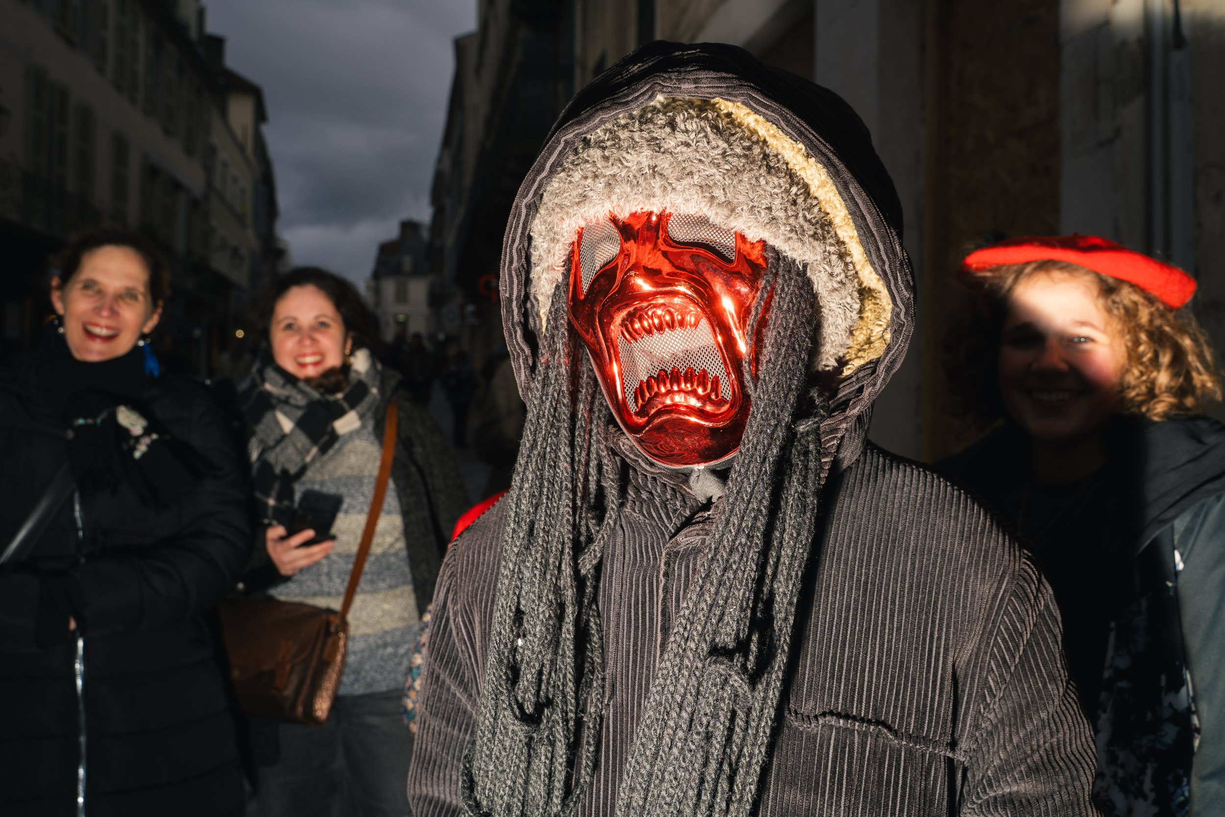A person wearing a red metallic mask with an aggressive expression, a hooded jacket, and dreadlocks on a city street at night, with three smiling women in the background.
