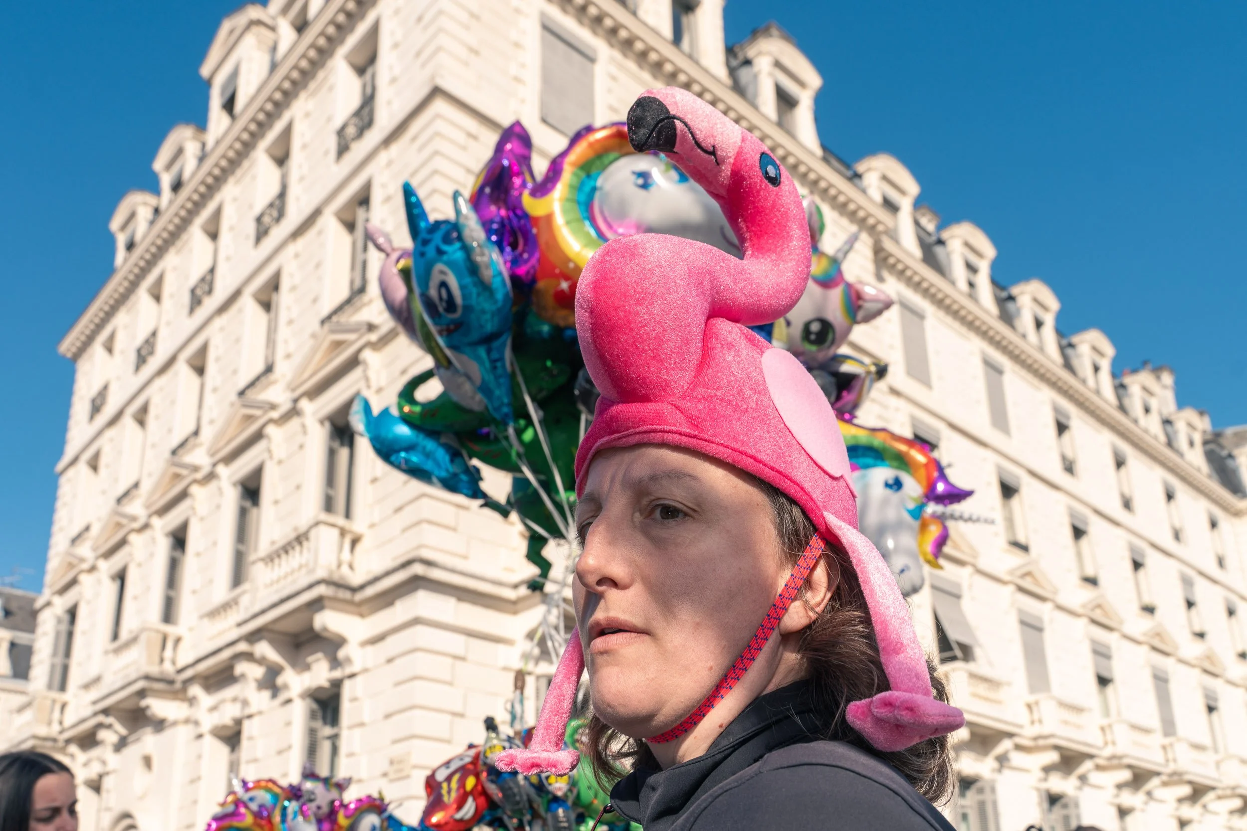 une femme porte un chapeau en forme de flamant rose qui se mélange avec des ballons à l'helium de toutes les couleurs sur le boulevard des Pyrénées à Pau pendant le carnaval Biarnès de Pau