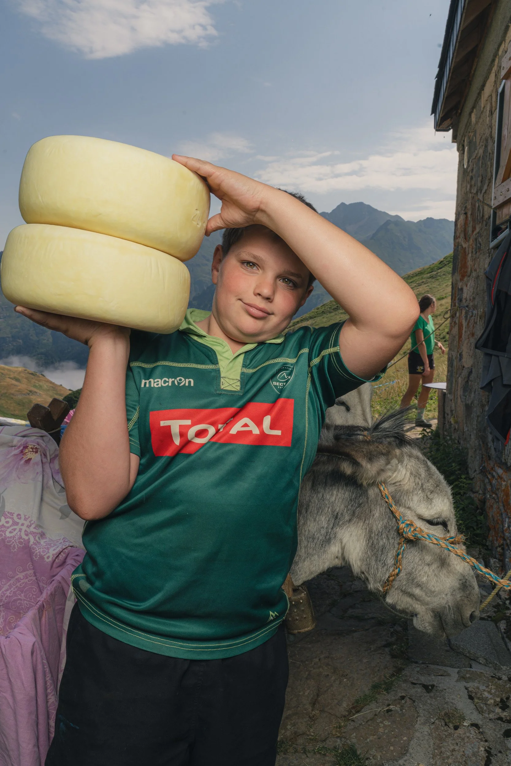 A boy holding two large wheels of cheese on his shoulder in a mountainous outdoor setting with a donkey standing nearby.
