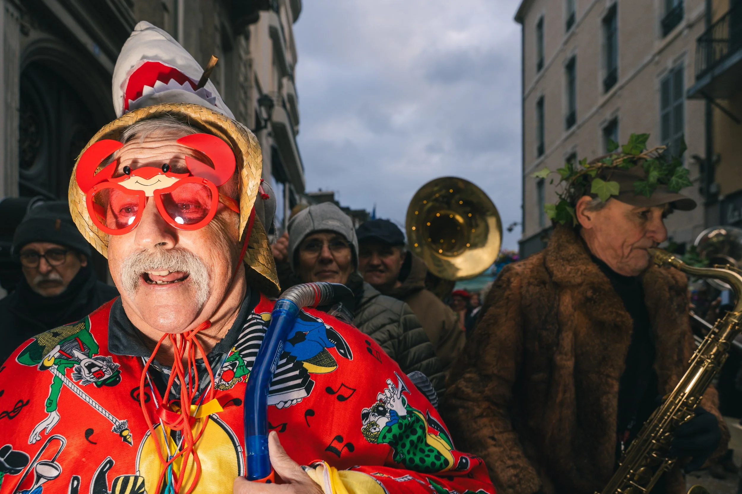 A man dressed in colorful costume with crab-shaped glasses and a straw hat decorated with a shark fin, standing among a crowd during an outdoor event or festival. Others in the background are playing musical instruments.