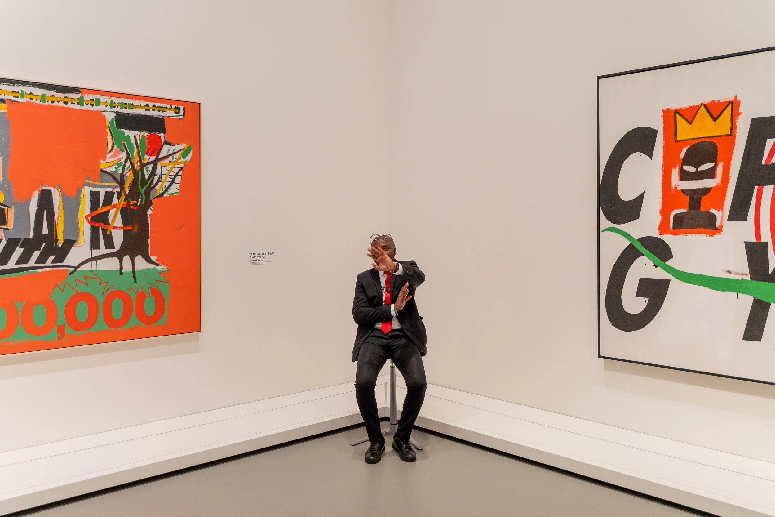 A man in a black suit with a red tie sits on a stool in an art gallery, covering his face with his right hand, with two large colorful abstract paintings on the white walls behind him.
