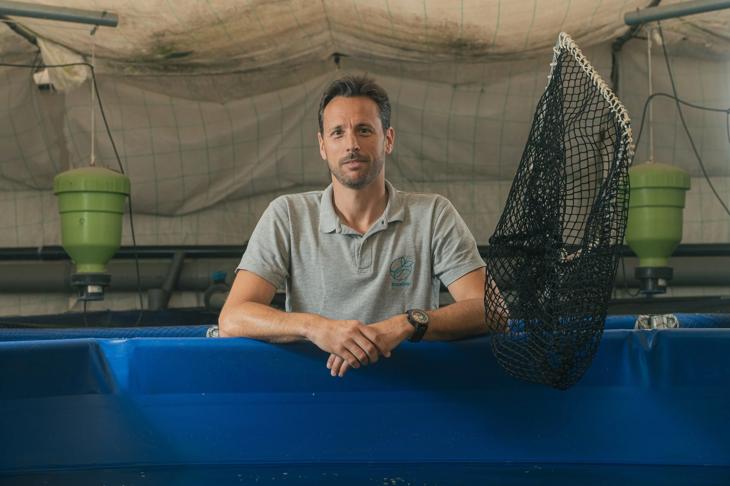Man in a gray polo shirt leaning on a blue tank with a black fish net hanging beside him in a room with netting on the ceiling and green water filtration devices.