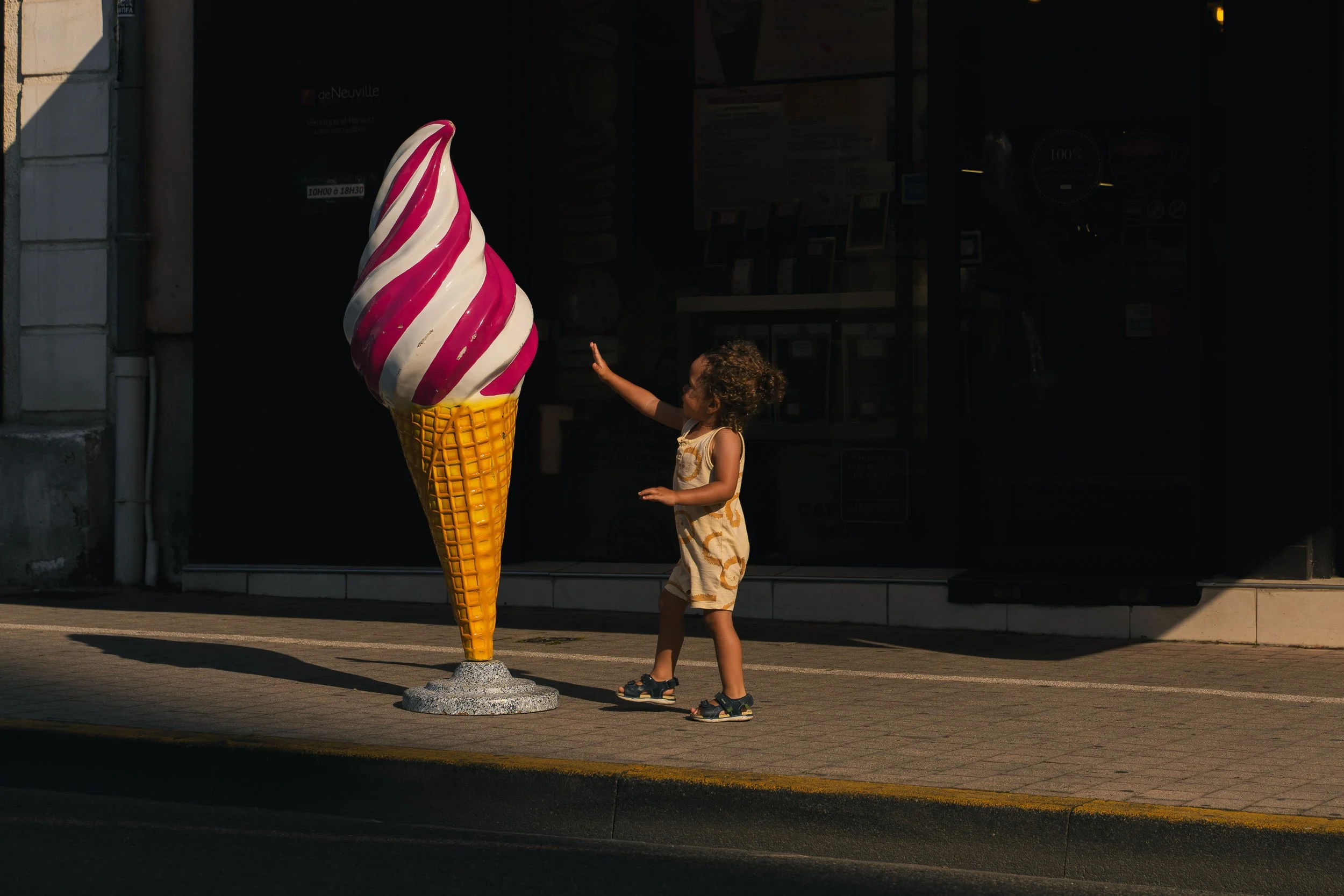 A young girl in a light-colored dress and sandals standing on a sidewalk, reaching out to a large ice cream cone statue outside a shop, with sunlight casting shadows.