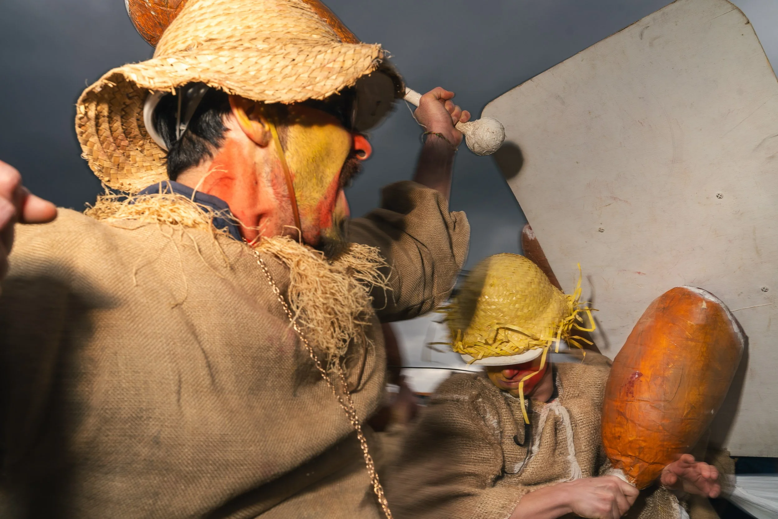 People dressed in traditional costumes, including straw hats and burlap clothing, appearing to play musical instruments or perform in a cultural setting.