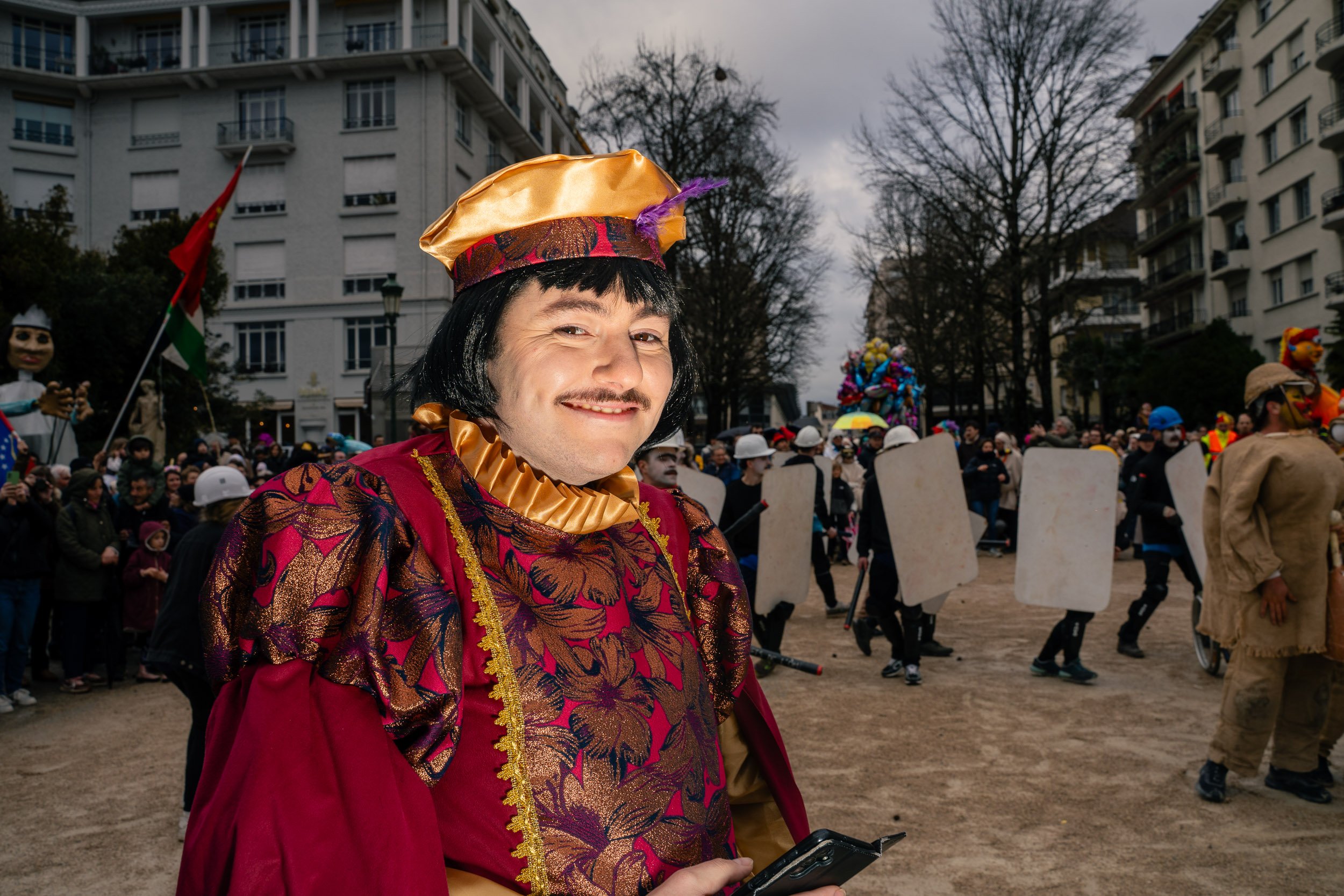 Person dressed in a colorful costume with a black wig and gold accessories at a parade with a crowd, balloons, and buildings in the background.