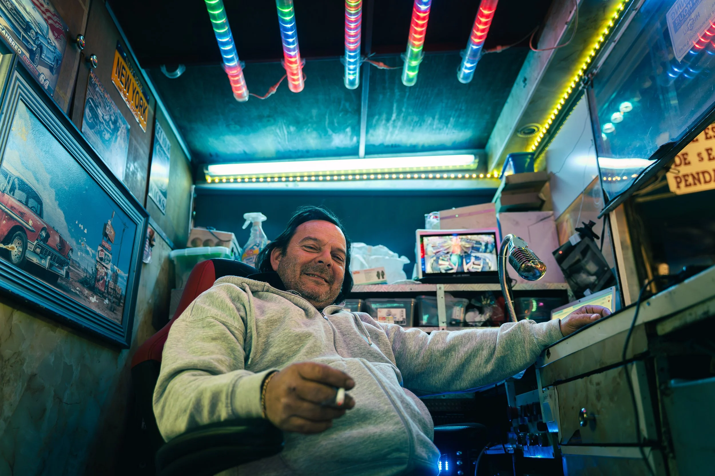 A man with shoulder-length dark hair smiling and sitting in a cluttered room with neon lights, surrounded by computer screens, posters, and various objects, holding a cigarette.