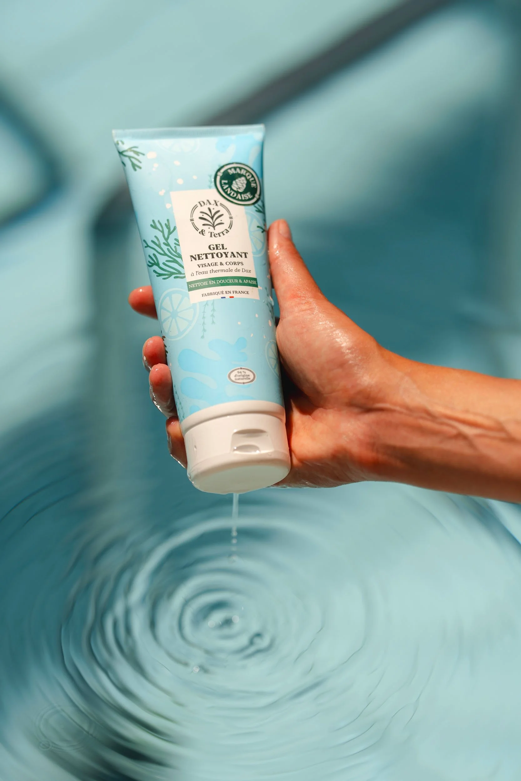 A person holds a tube of gel cleanser over a light blue water-filled basin, with water droplets on their hand.
