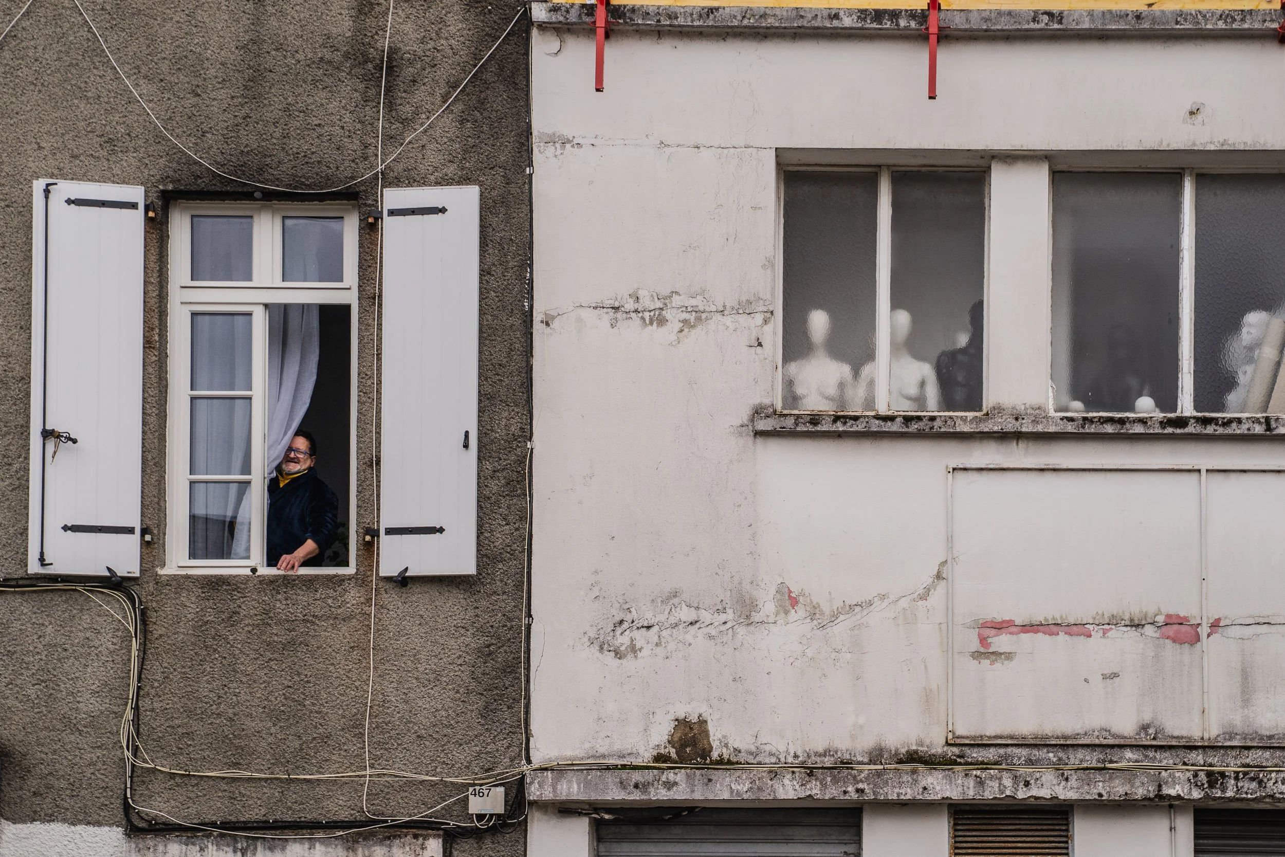 A man looking out of an open window with white shutters in an older, weathered building with peeling paint and cracks, and a row of mannequin heads visible through the foggy window on the adjacent building