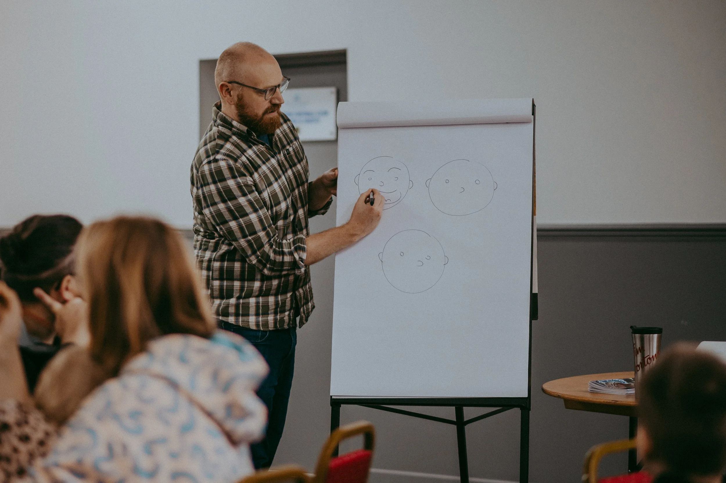 A man with glasses and a beard wearing a plaid shirt stands at a whiteboard drawing cartoon faces of a happy, a neutral, and a sad face during a presentation in a classroom or conference room.