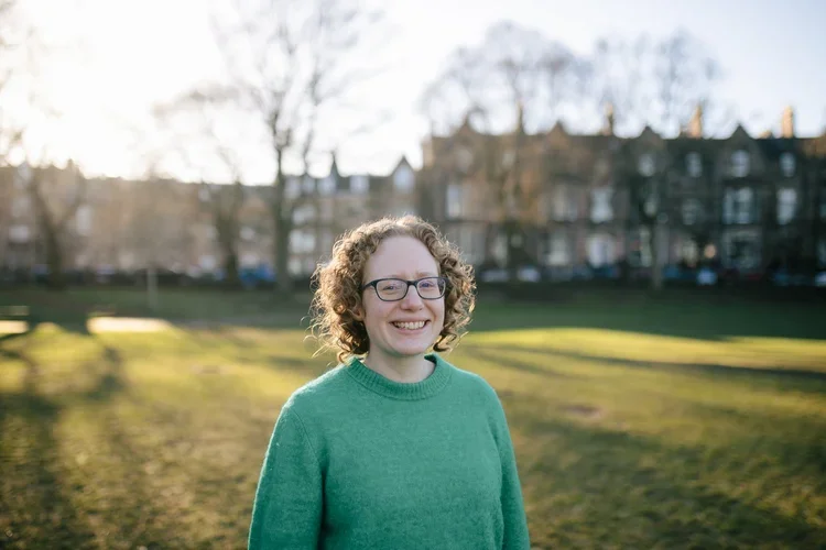 A fairskinned woman with curly hair and glasses smiling outdoors in a park on a sunny day.