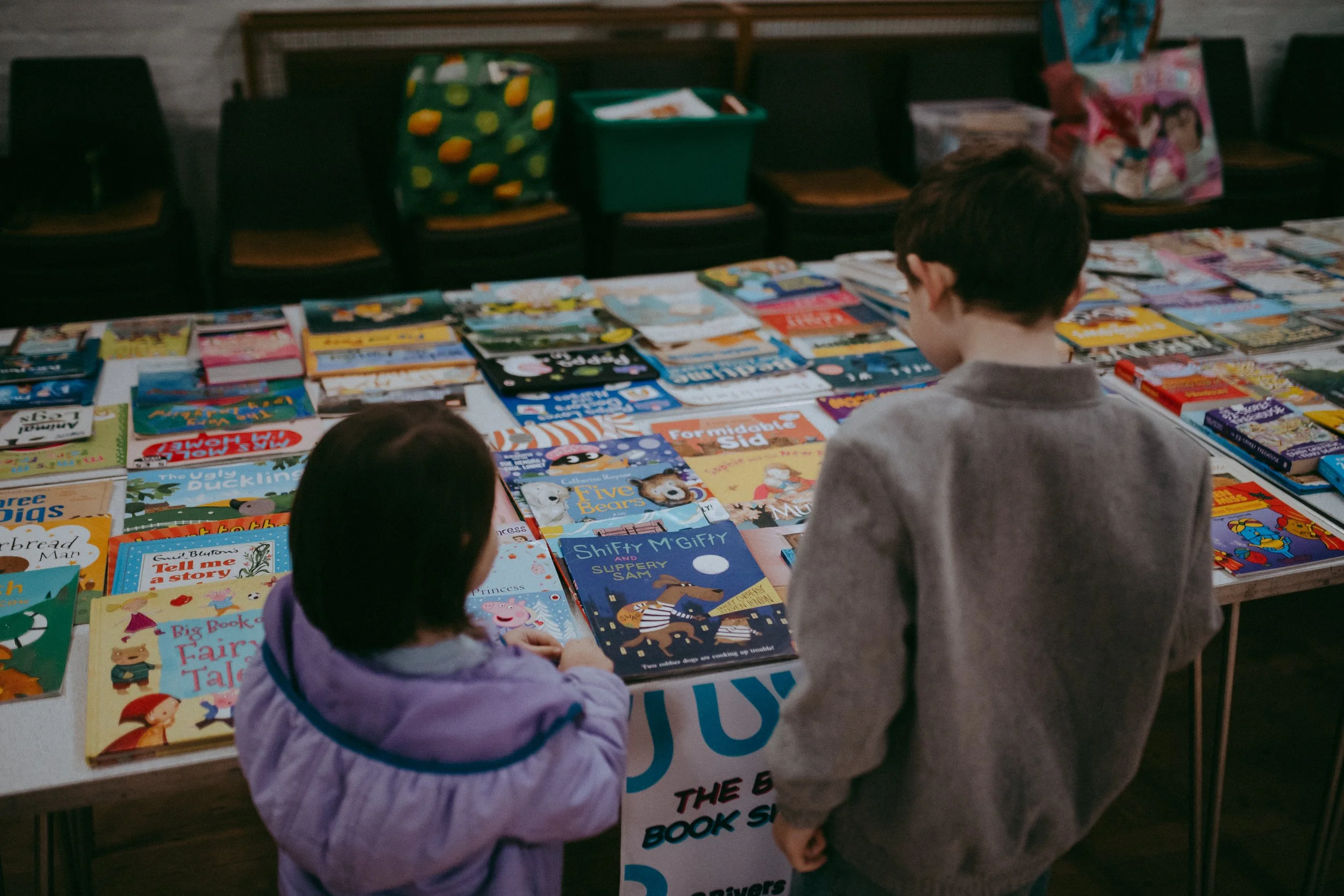Two children browsing through children's books on a table at a book sale or library event.