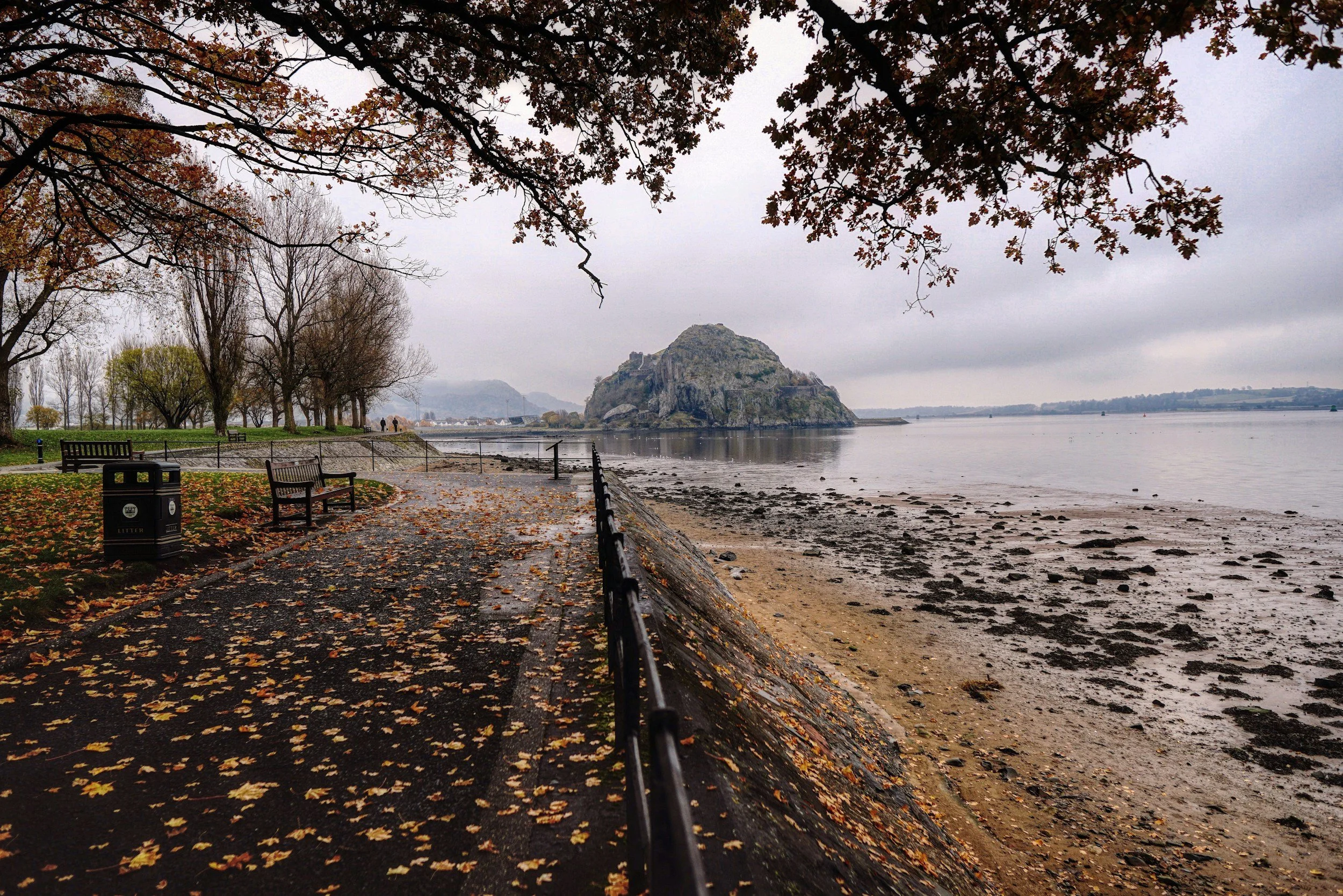 A riverside park with benches, leaf-covered walkway, and trees with autumn leaves overlooking a calm body of water. A large rocky island - Dumbarton Rock - is in the water, with cloudy skies above.