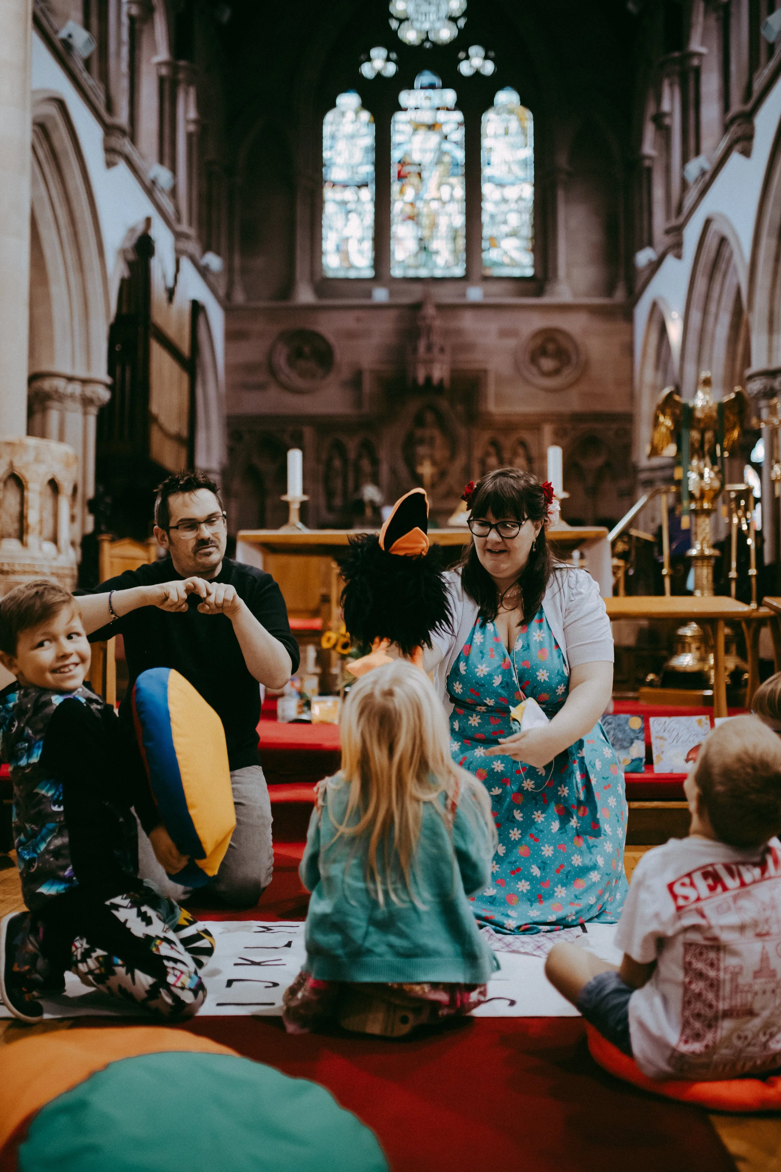 Children and adults sitting on the floor inside a church during a storytelling event, with stained glass windows and wood paneling in the background.