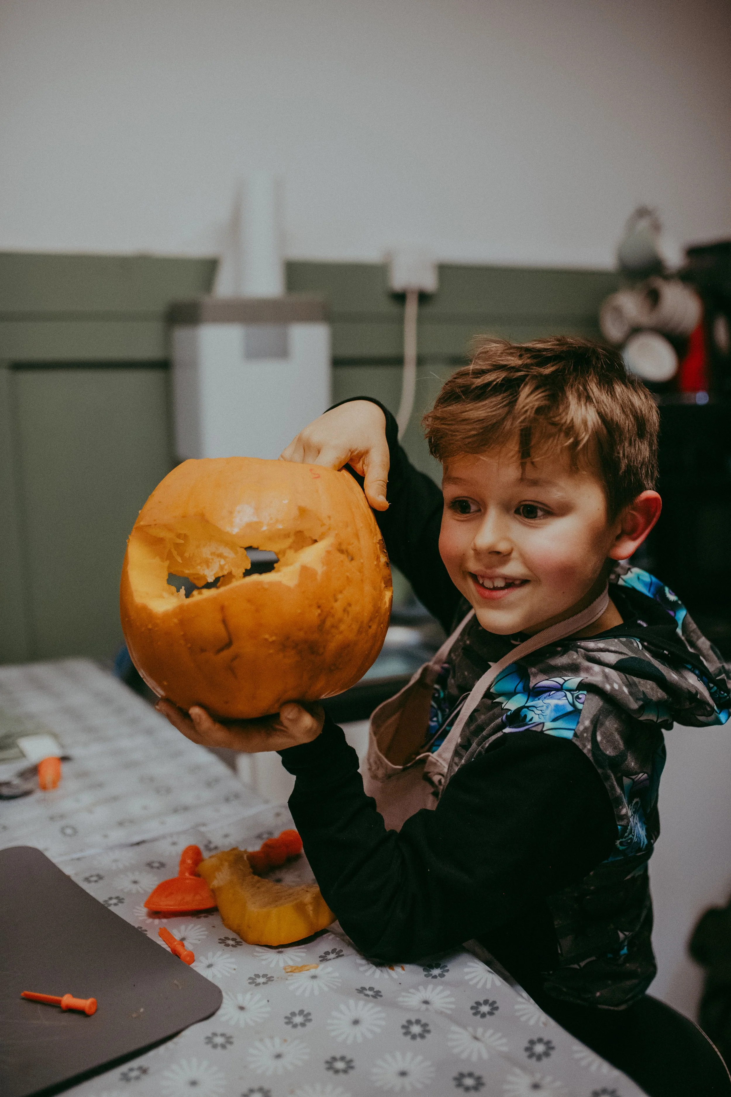 A young boy with brown hair smiling and holding a carved pumpkin with a face design. He is sitting at a table covered with a festive tablecloth, with pumpkin carving tools and pumpkin pieces on the table. In the background, there are kitchen applianc