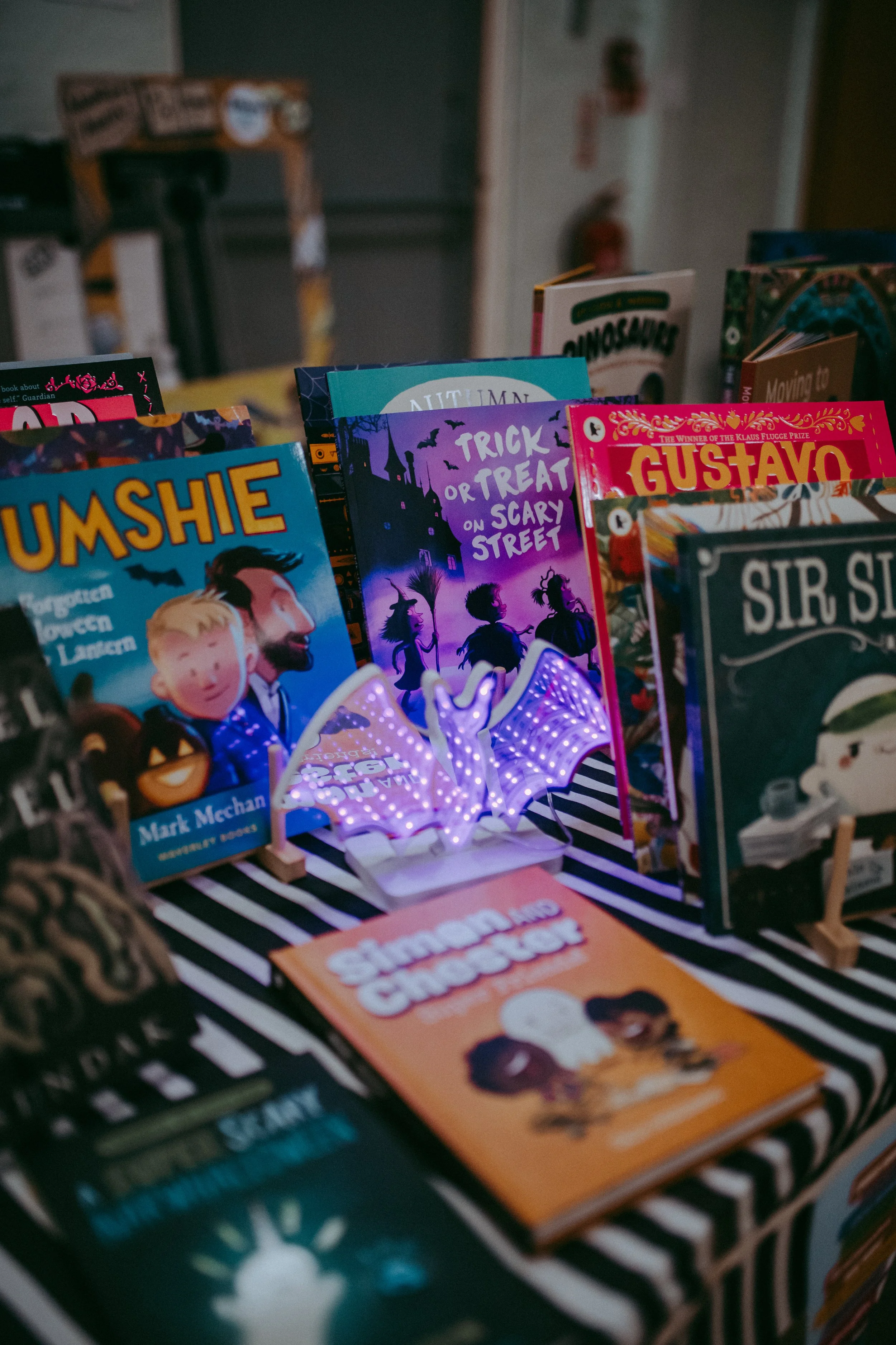 A bookshelf with children's books, including titles like 'Tumshie', 'Trick or Treat on Scary Street', and 'Gustavo'. There is a purple light-up bat decoration in front of the books and a striped black and white tablecloth underneath.