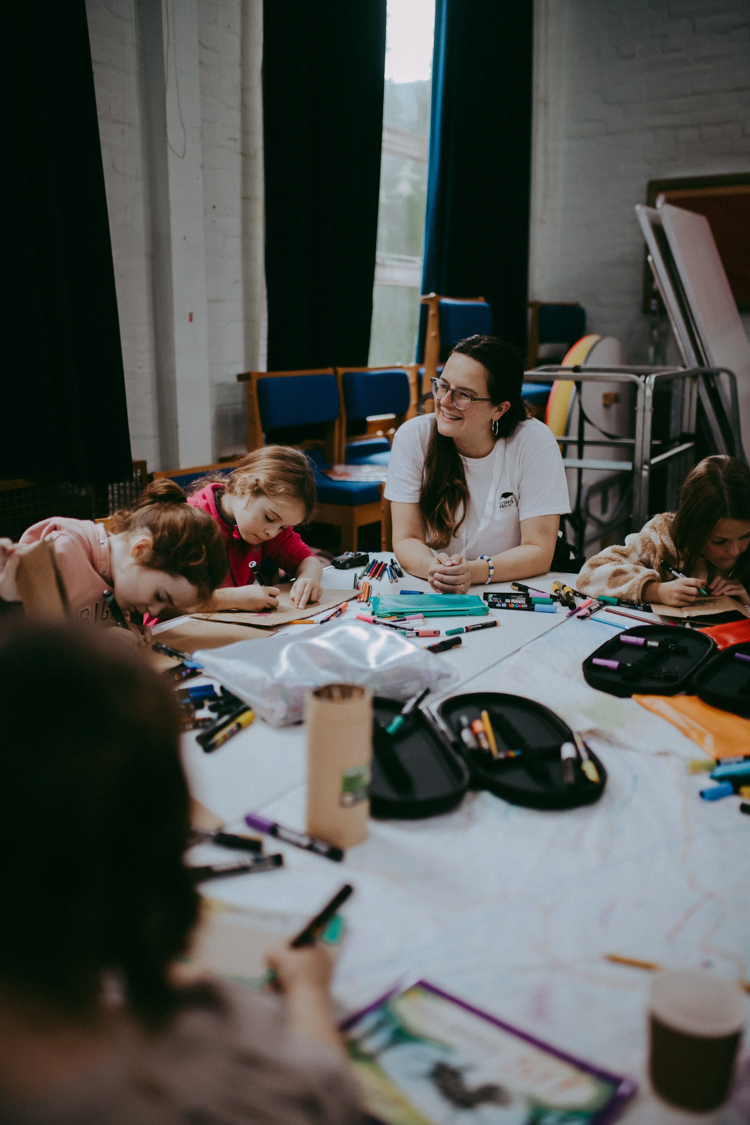 A woman with glasses smiling while working with children at a table cluttered with art supplies, in a room with chairs and windows.