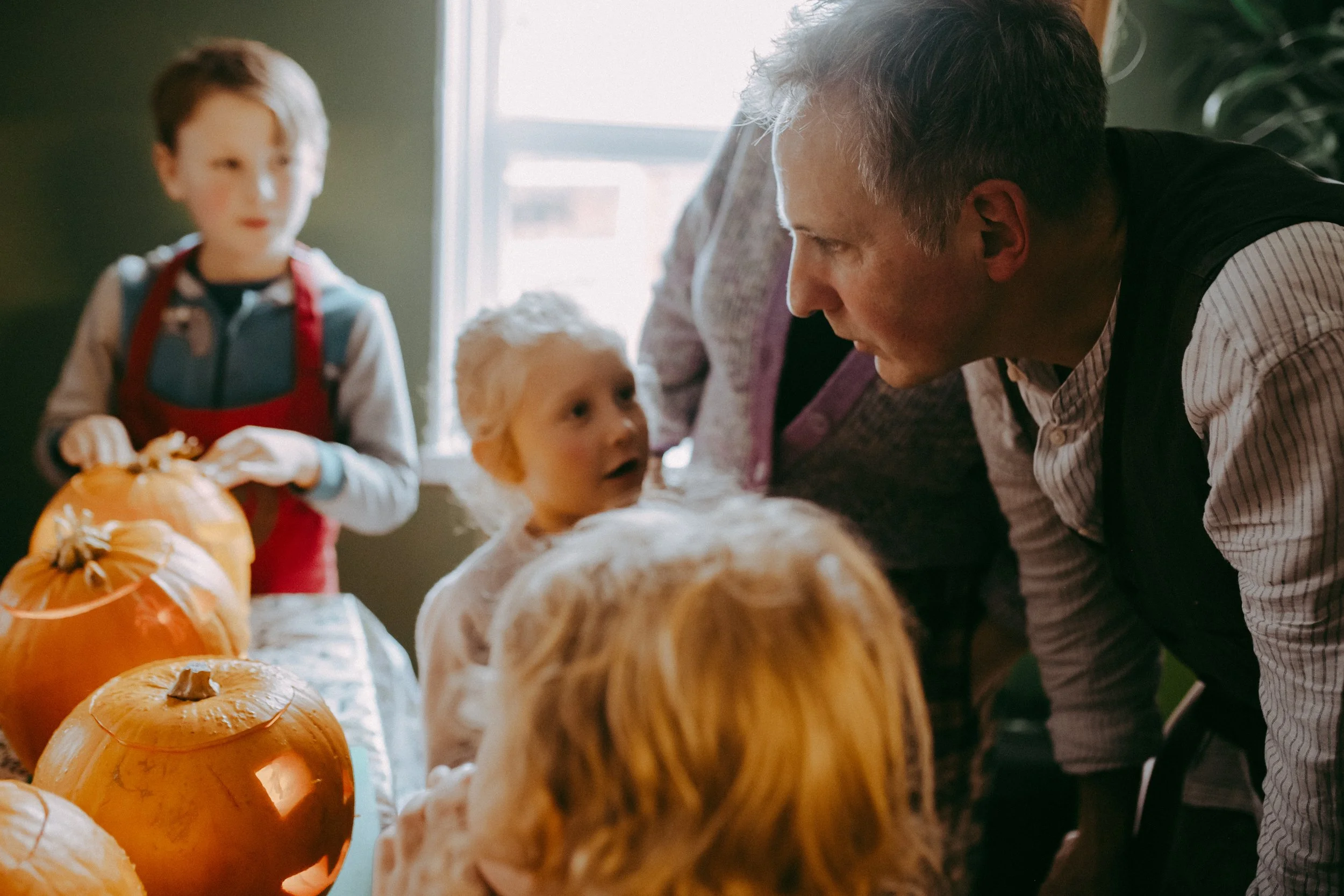 Family gathering around pumpkins, possibly preparing for Halloween or fall celebration, with children and an adult in a cozy indoor setting.
