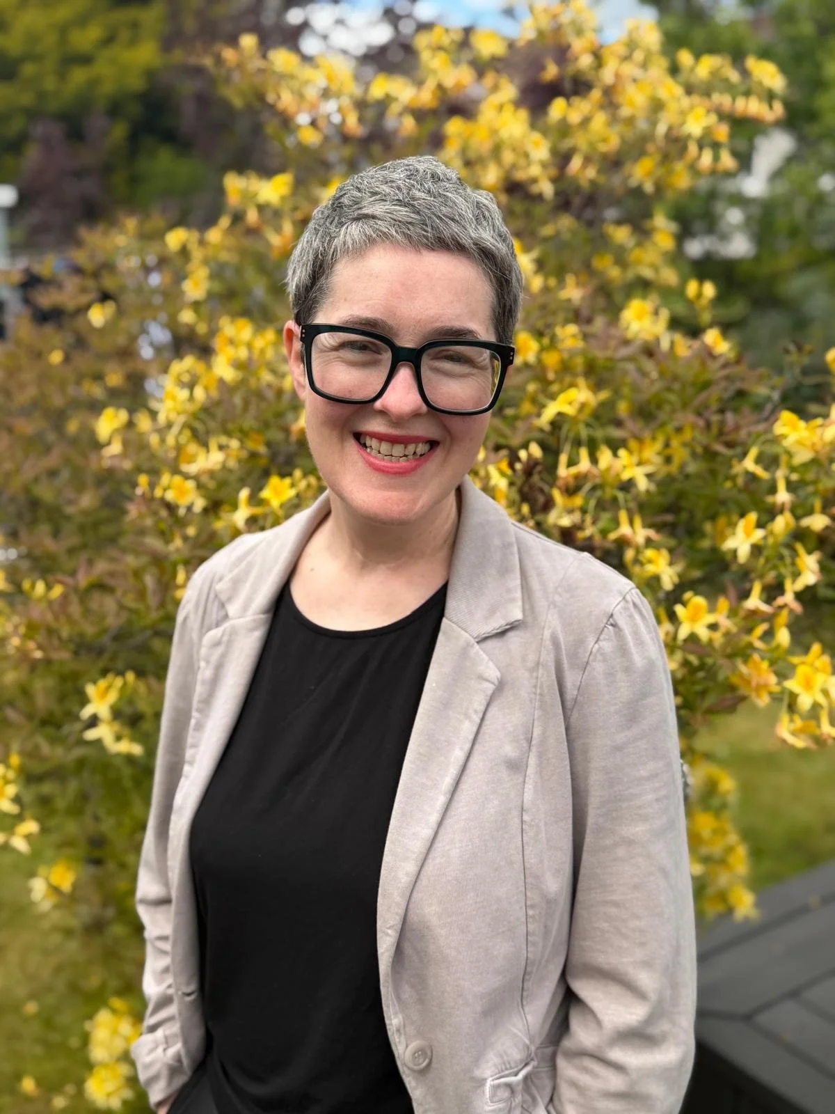 A smiling woman with short gray hair and large black glasses, wearing a beige jacket and black shirt, standing outdoors in front of yellow flowering bushes.