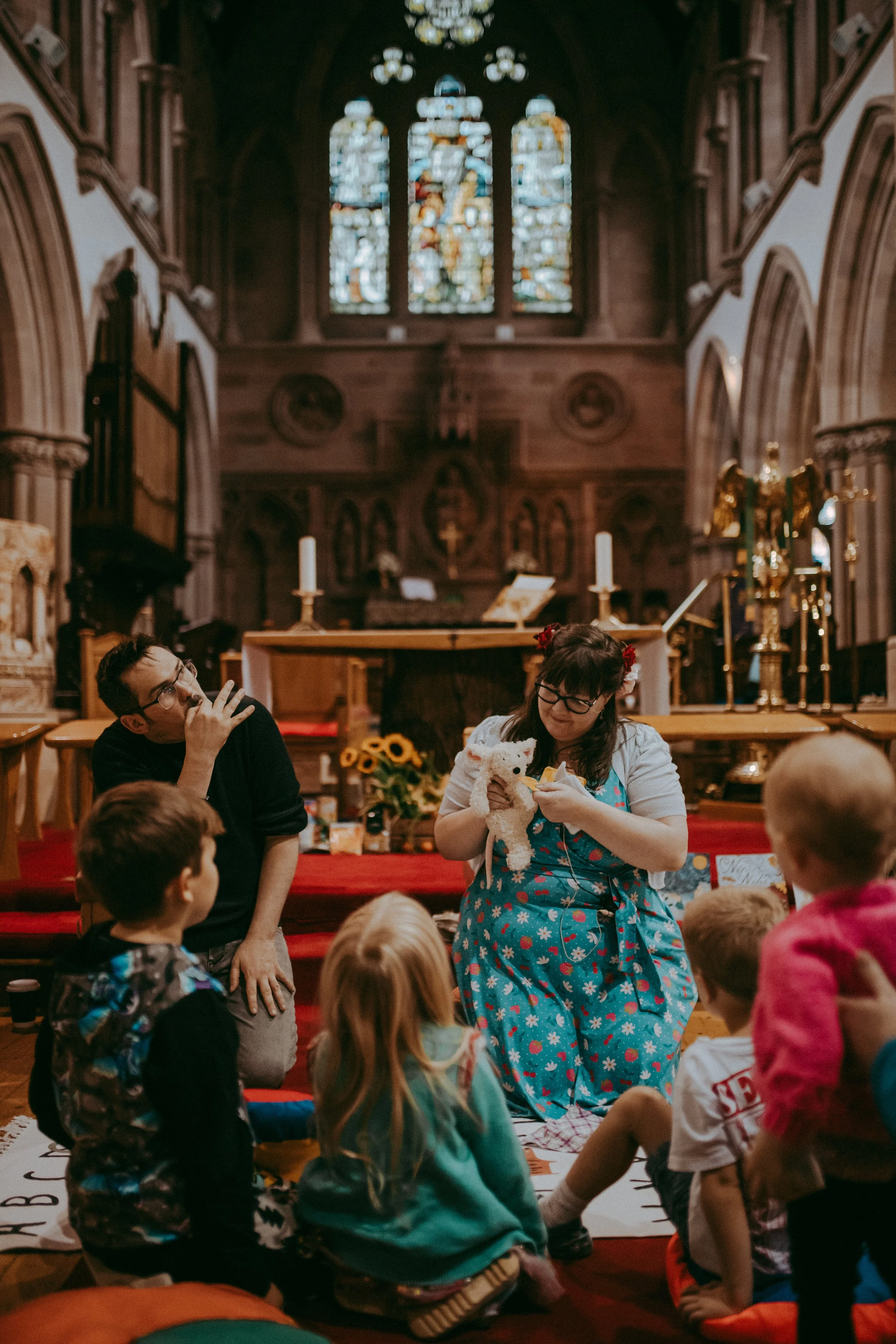 Inside a church with stained glass windows, children sit on a red carpeted floor listening to a woman who is sitting and holding a stuffed animal. Two adults are present. The scene appears to be a storytelling or educational session for children, wit