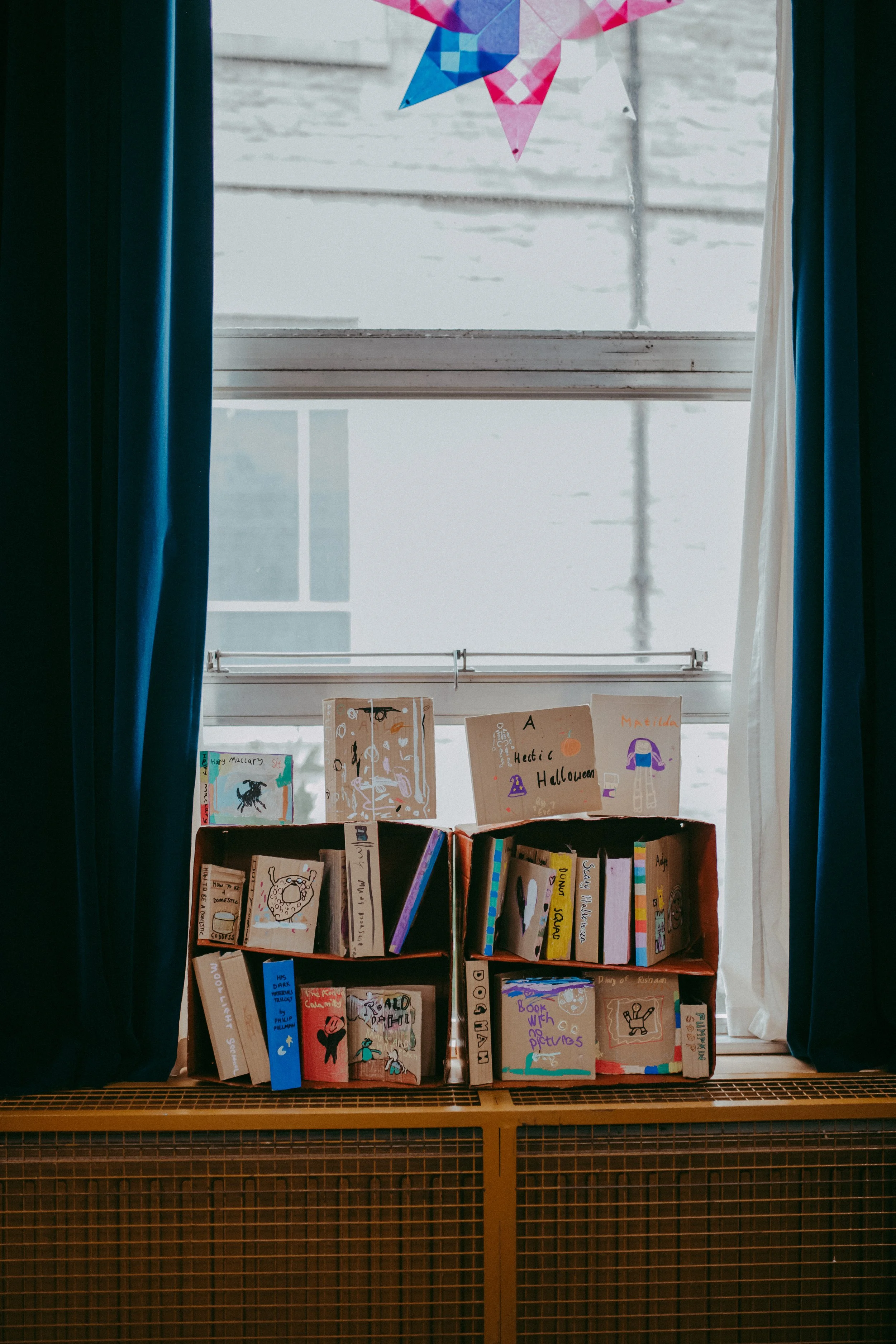 Books displayed on a wooden shelf underneath a window with colorful artwork hanging above, and blue curtains on each side.