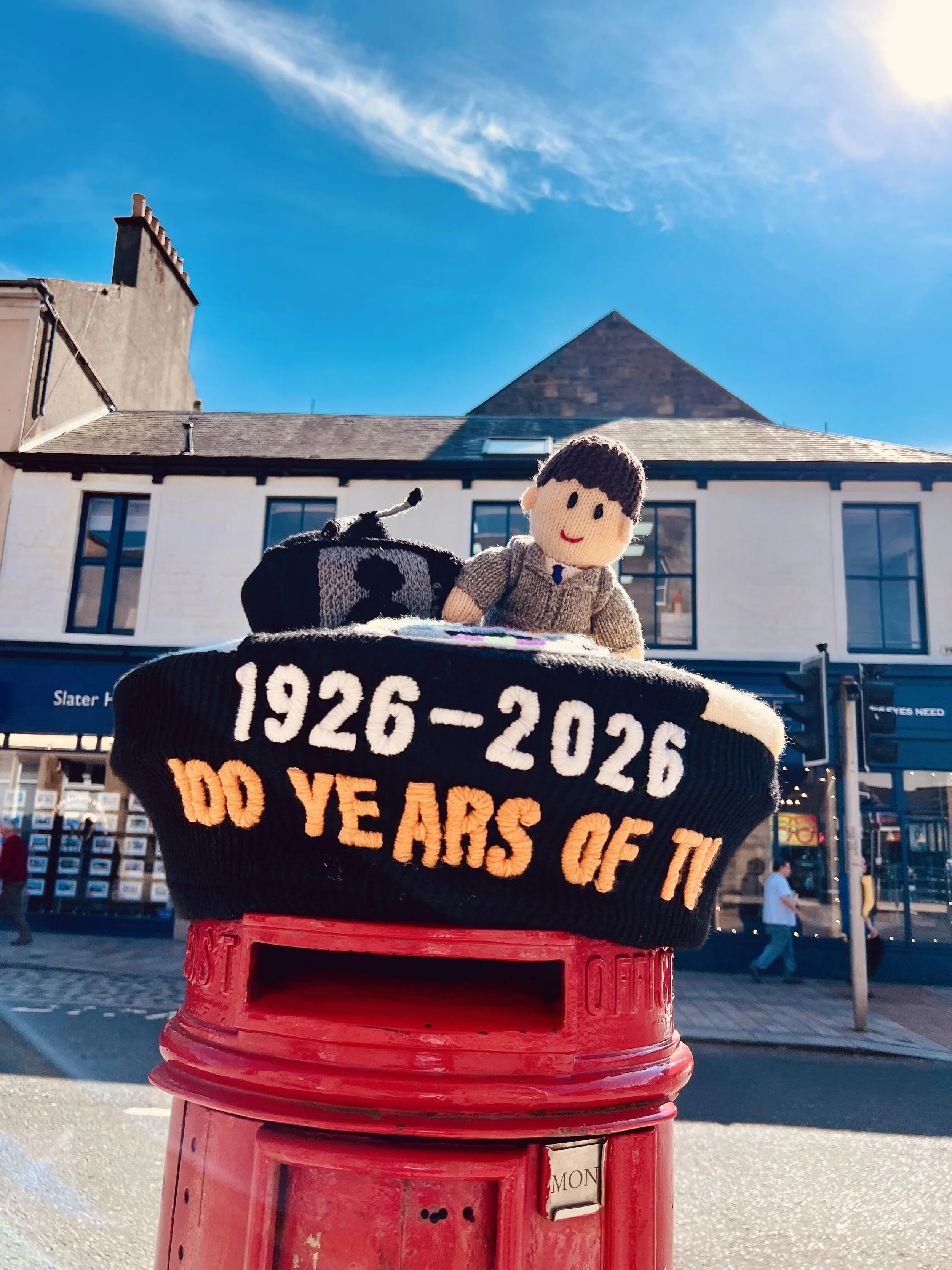 A red mailbox with a black knitted cover featuring a smiling doll figure and the text "1926-2026" and "50 YEARS OF T". The mailbox is topped with a knitted doll, and the background shows a building with white walls and blue windows beneath a bright blue sky with a few clouds.