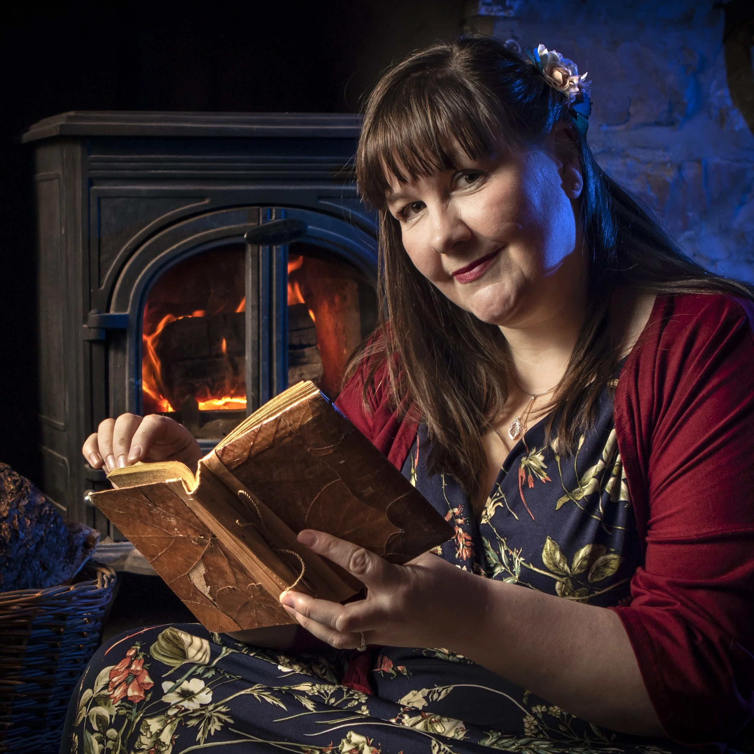 A fair skinned woman with long brown hair, wearing a floral dress and a red jacket, is sitting near a fireplace reading a book.