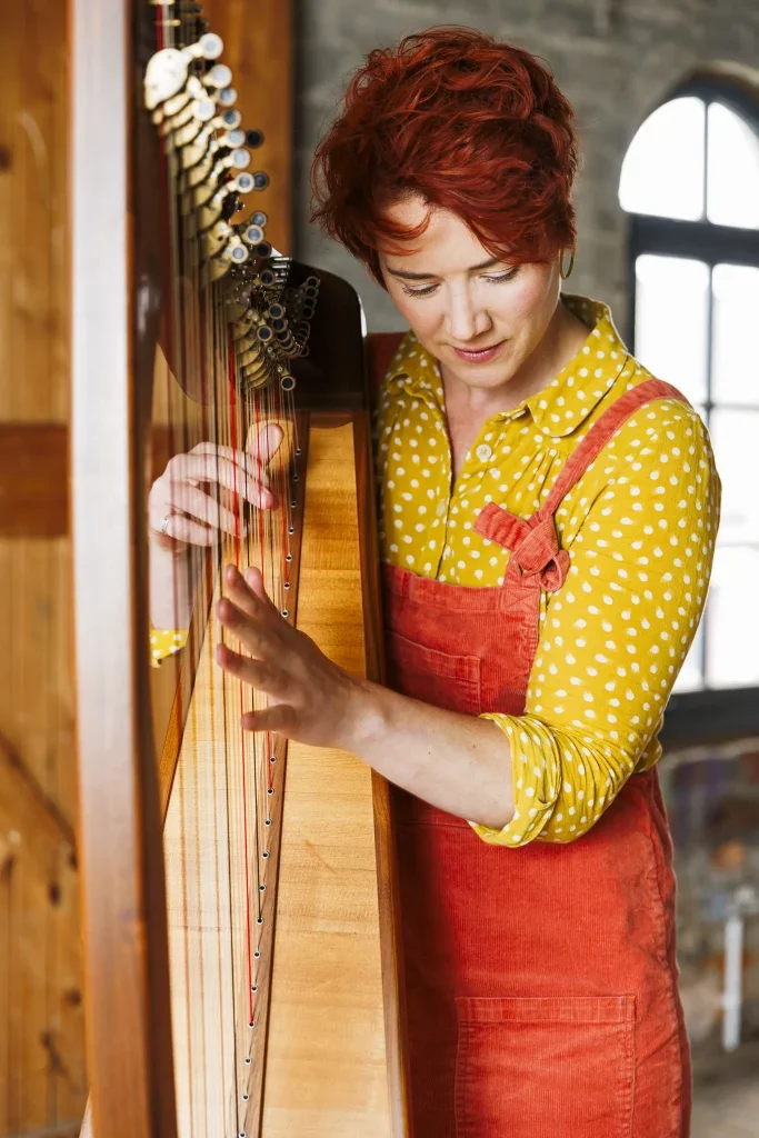 A fair skinned woman with short red hair and a yellow polka dot blouse is playing a wooden harp, looking down at the strings.