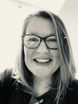 A fair skinned woman with light-colored hair wearing glasses, smiling at the camera.