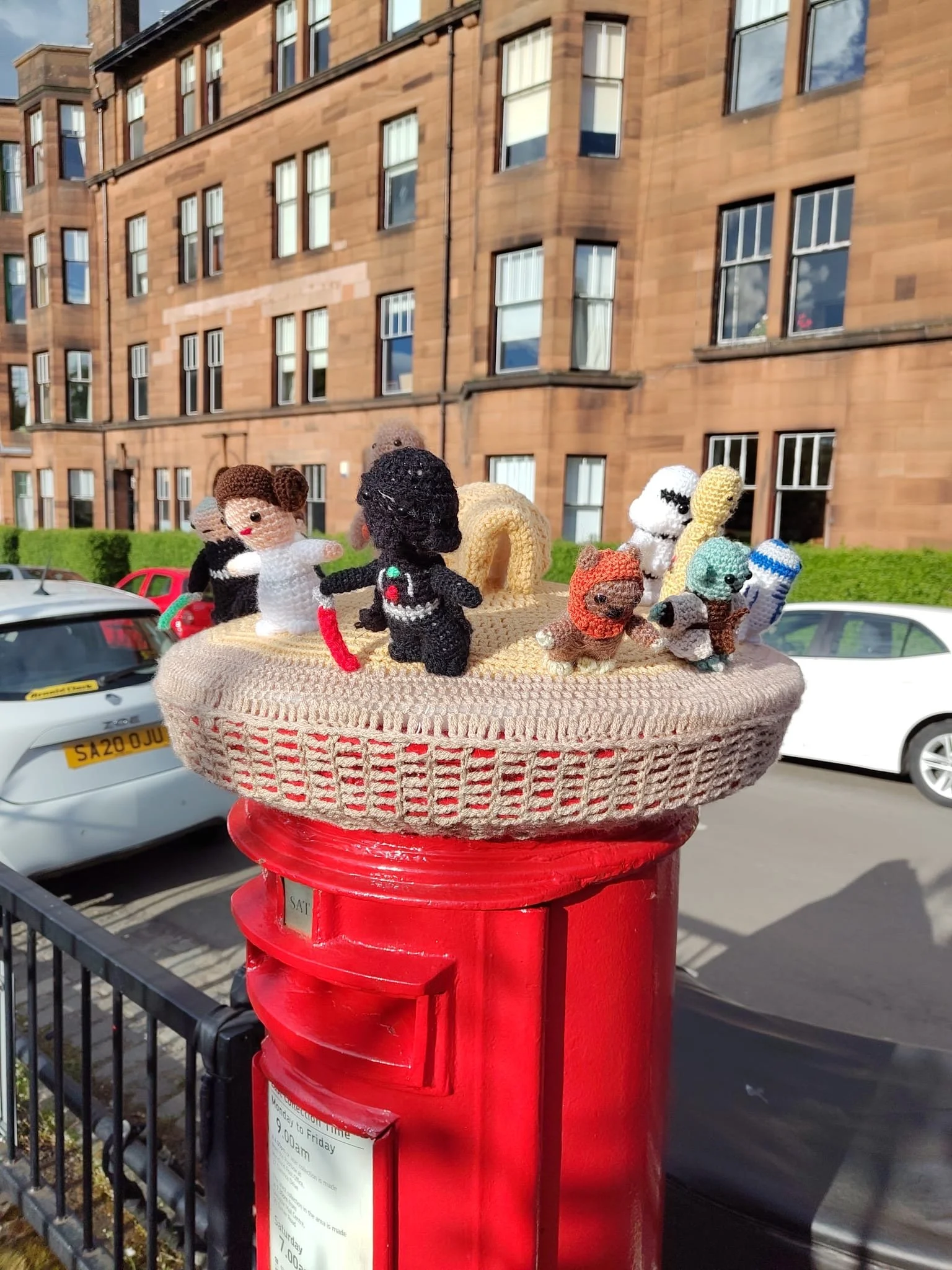 Small knitted stuffed animals placed on top of a red postbox in a city street, with cars parked nearby and a brick building in the background.