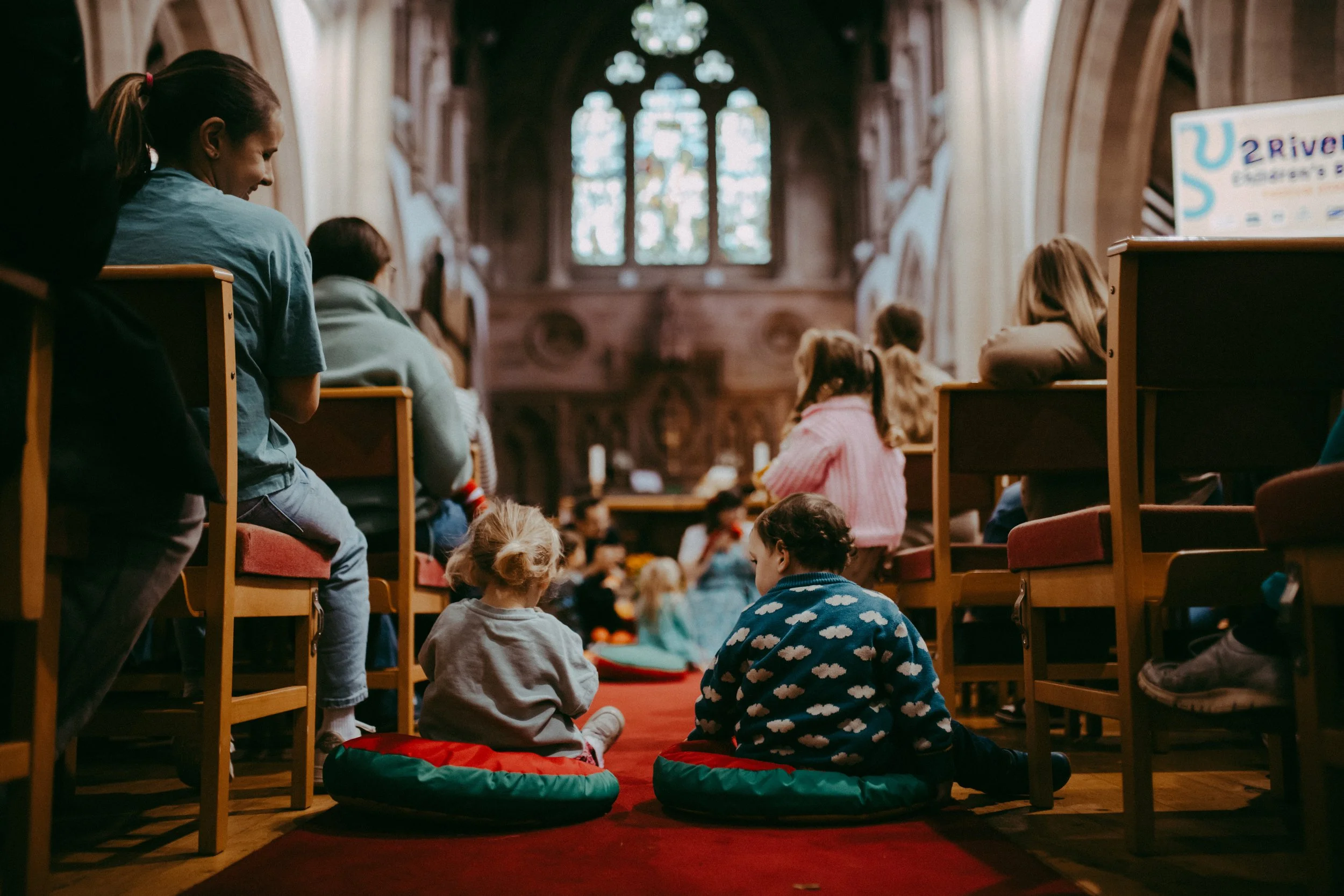 Children sitting and kneeling on a red carpet inside a church, facing the altar and a large stained glass window, during a storytelling event.