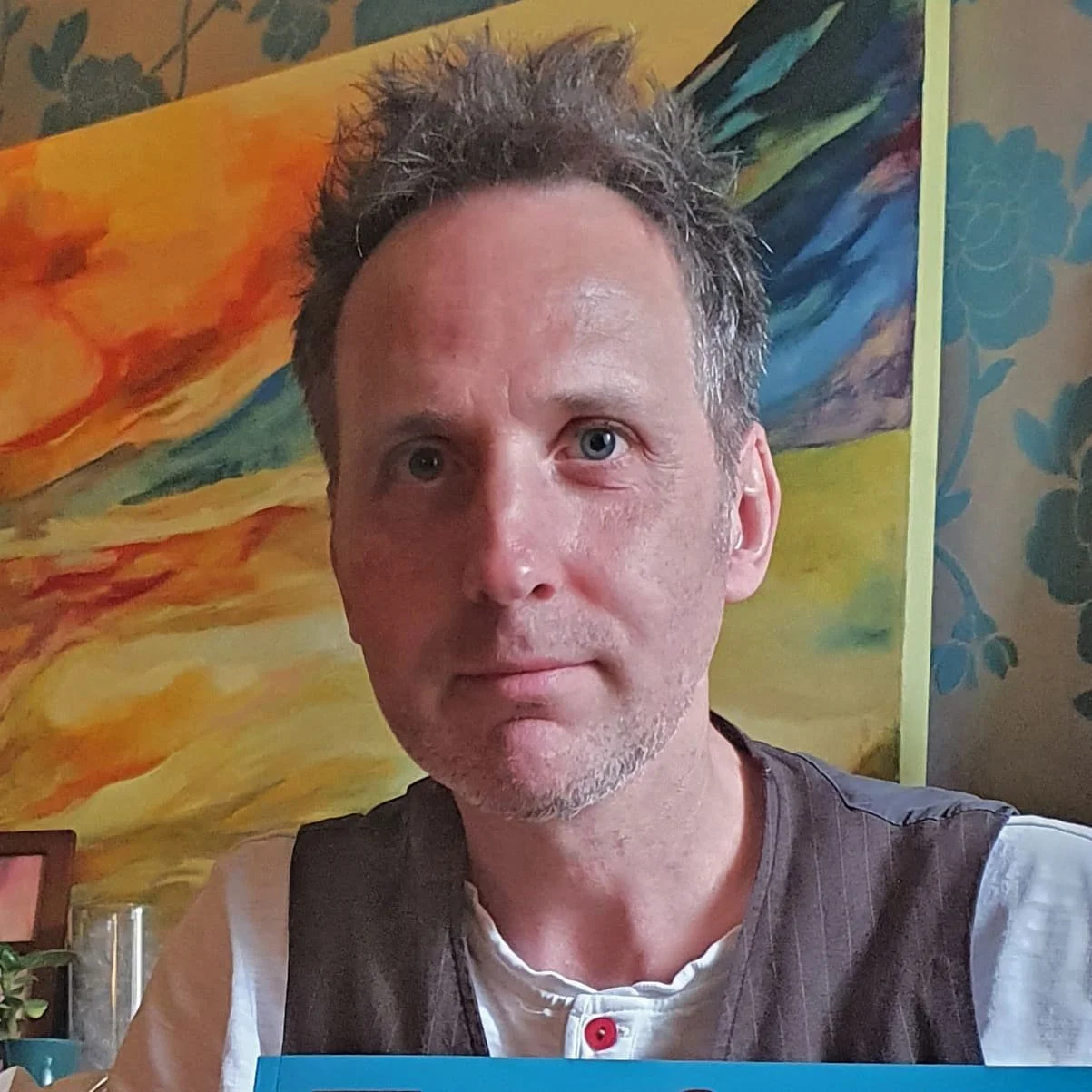 A fair skinned man taking a selfie indoors with colorful abstract art and floral wallpaper in the background.