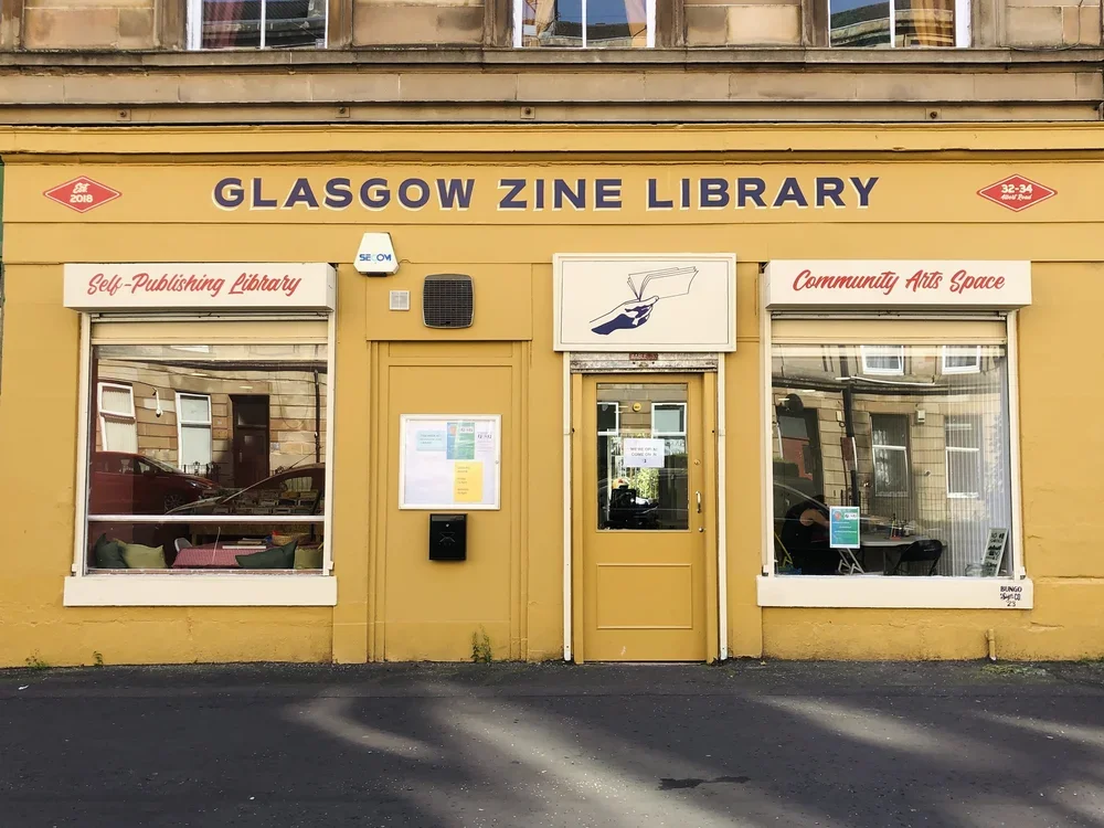 The exterior of a vibrant yellow building housing the Glasgow Zine Library, with signs indicating it's a self-publishing library and community arts space. The building has two large windows showing interior furnishings and a central door, with a sign above featuring an illustration of a hand holding a pen.