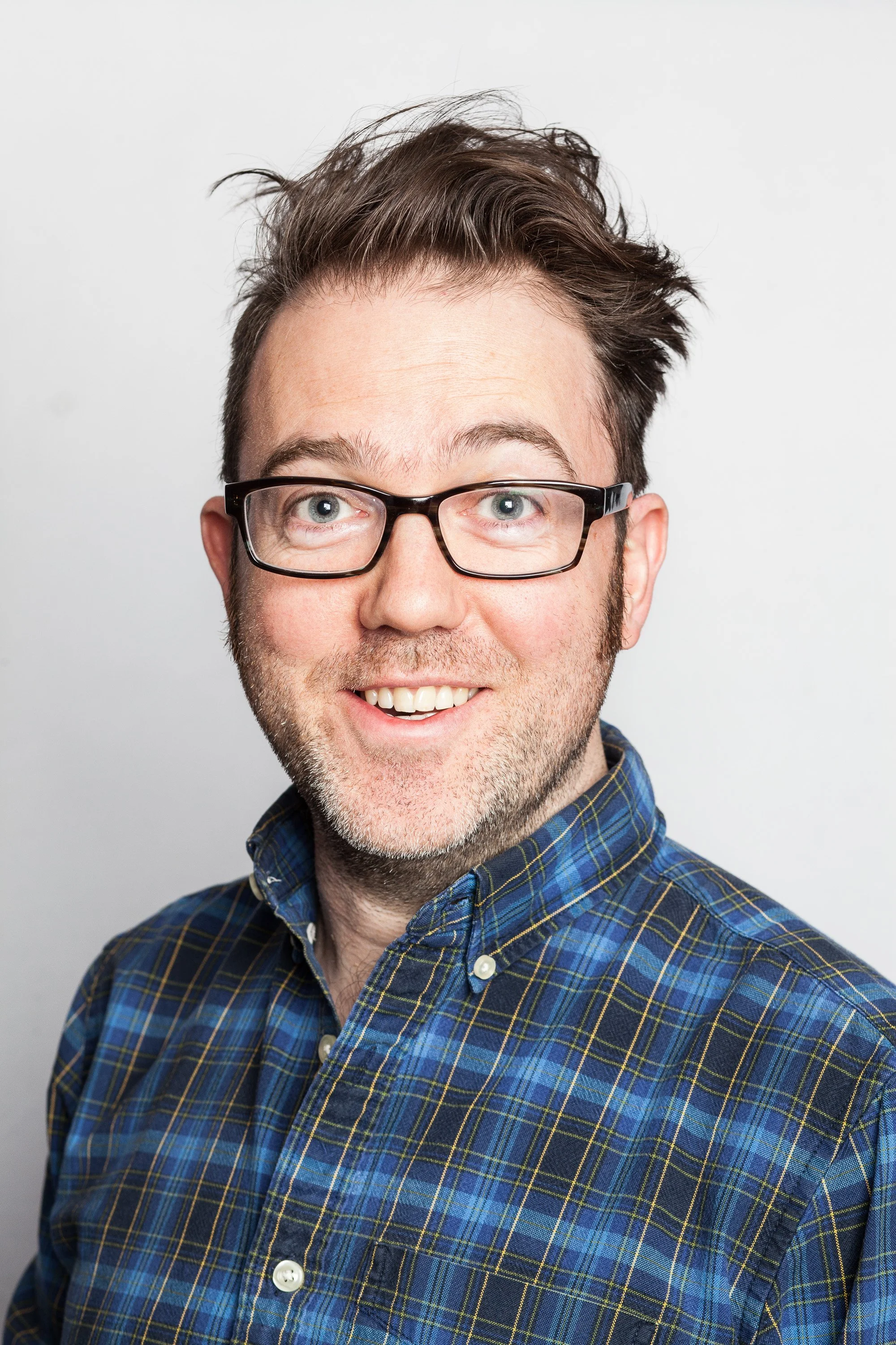 A fair skinned man smiling, wearing glasses, a blue plaid button-up shirt, with a white background.